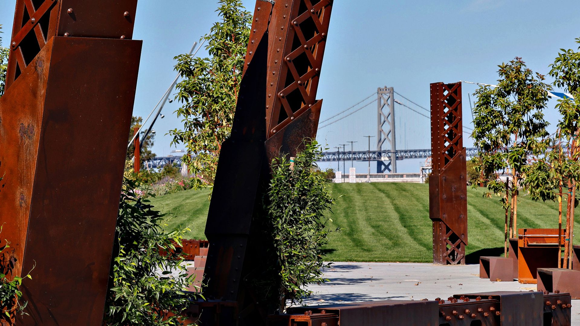 Photo of rusted, red steel beams in a grassy park with a bridge in the backdrop