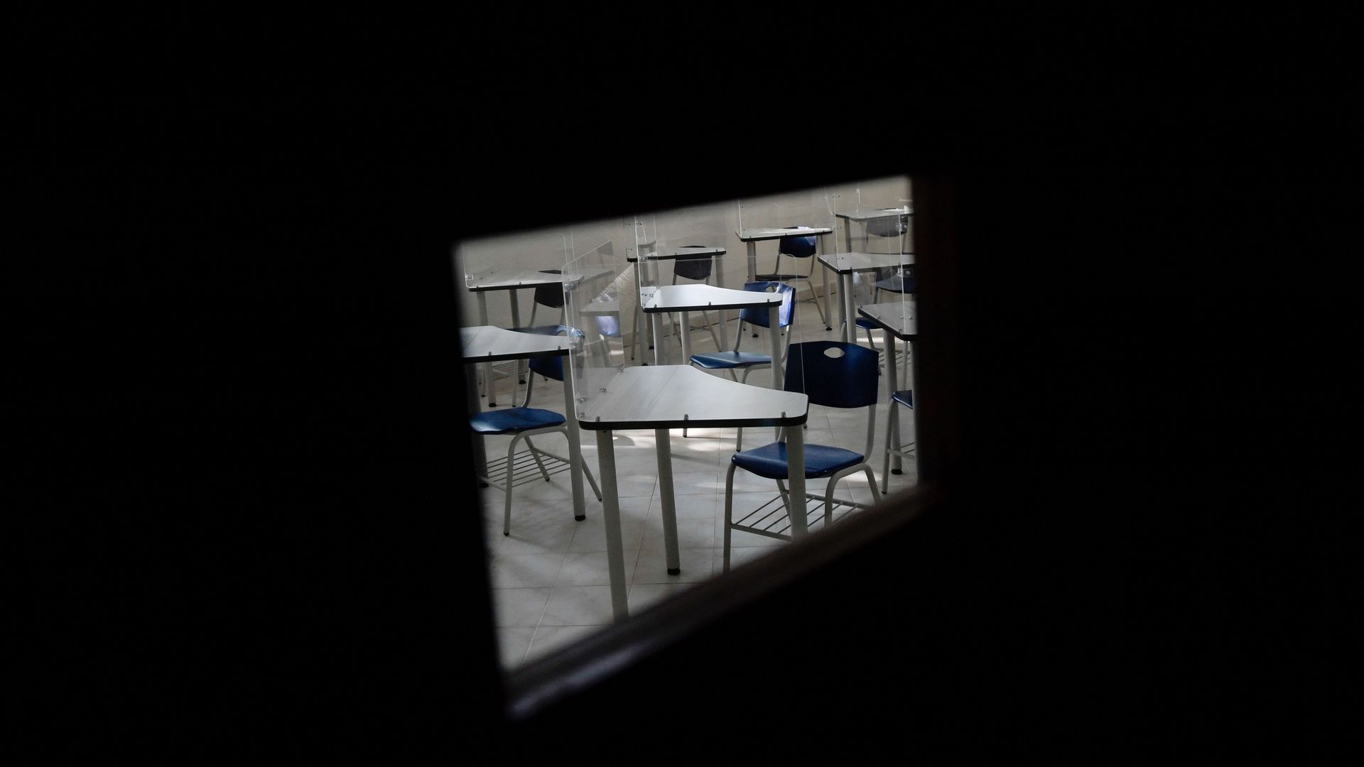 Empty desks in a classroom are photographed through a small window in a door.