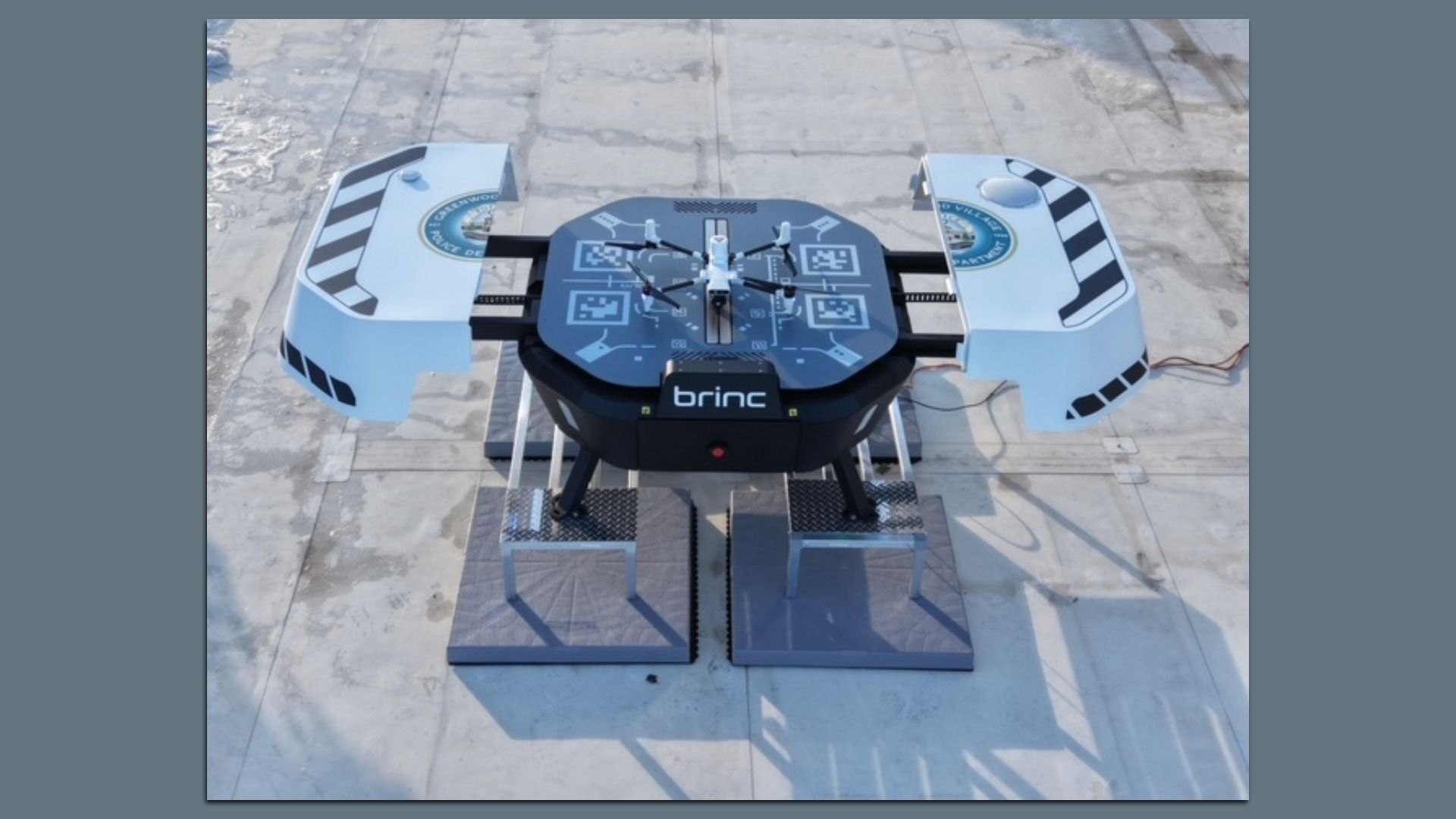 Quadcopter drone rests on a blue circular Brinc testing platform with QR-like markings; side panels show black-and-white stripes, all on a concrete outdoor pad.