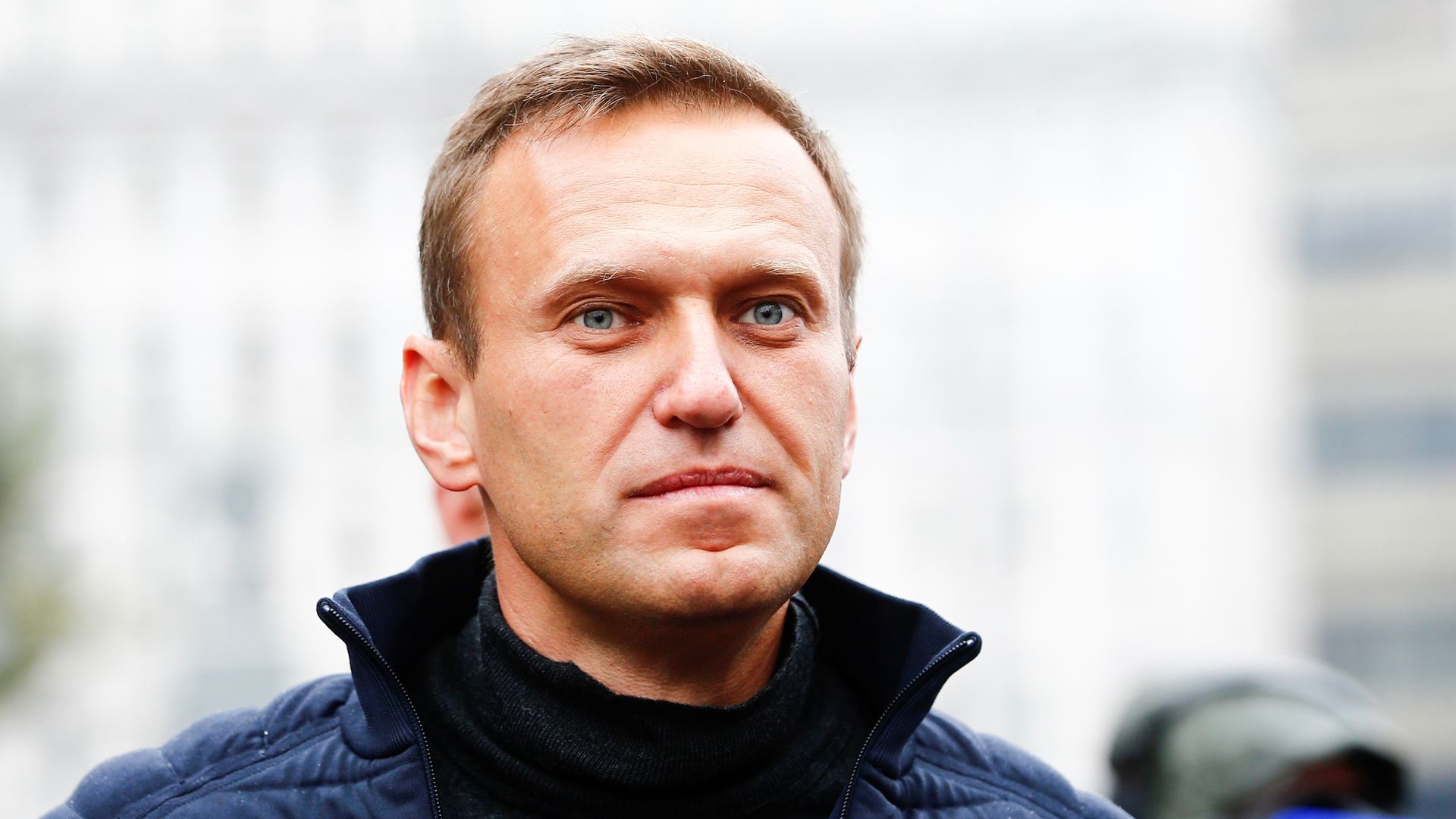 Alexei Navalny standing outdoors wearing a black jacket. The photo is a closeup of his face. 