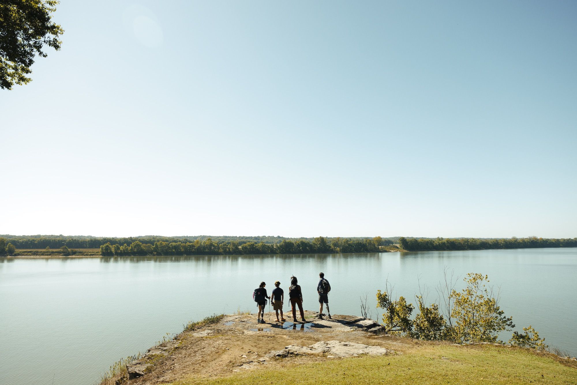 Four people standing on a mound overlooking a River, treeline and open sky.