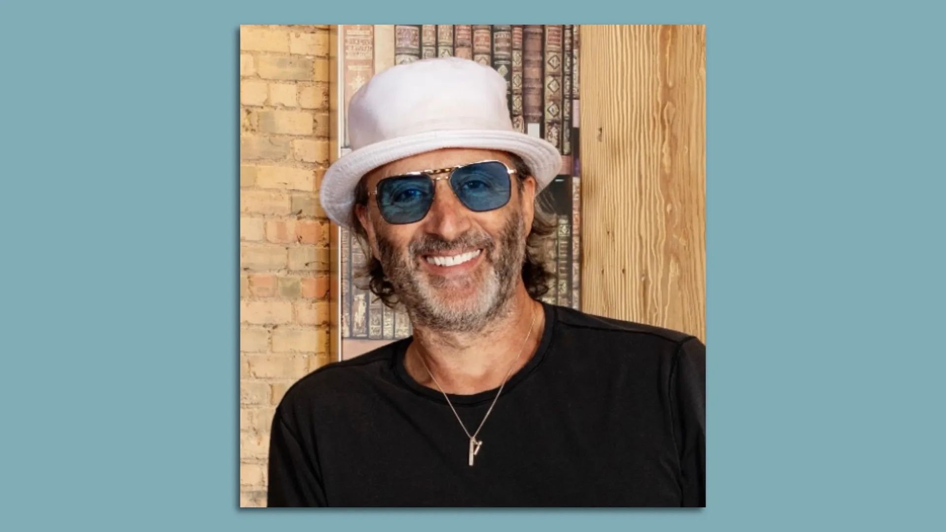 Smiling man with stubble wearing a white hat, blue sunglasses, black shirt, and pendant necklace stands in front of a wall with a book-patterned background and wood panel.