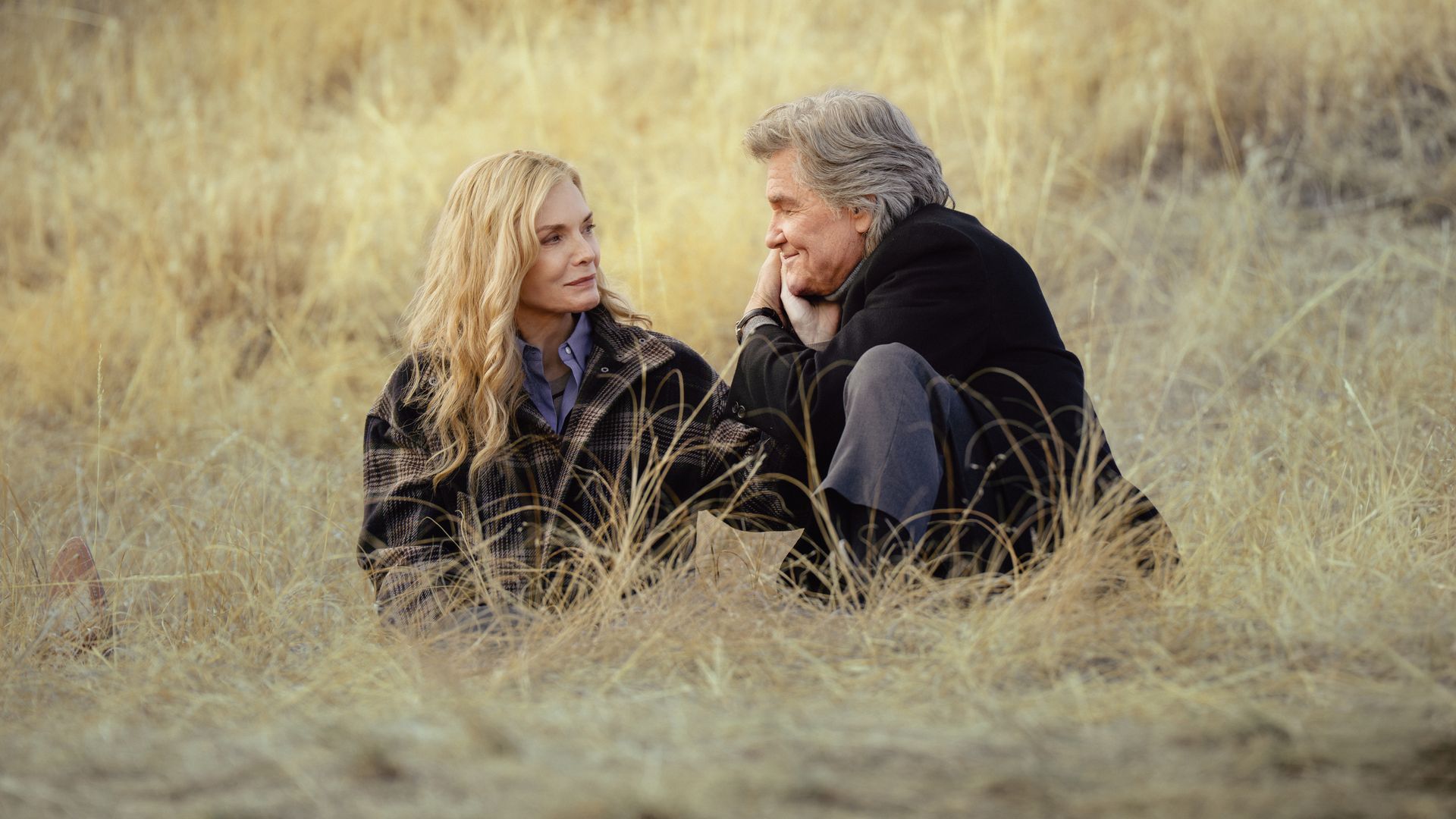 Two older adults sit in a sunlit golden meadow, facing each other. The blonde woman in a plaid jacket and the gray-haired man in a black coat share a quiet, warm moment.