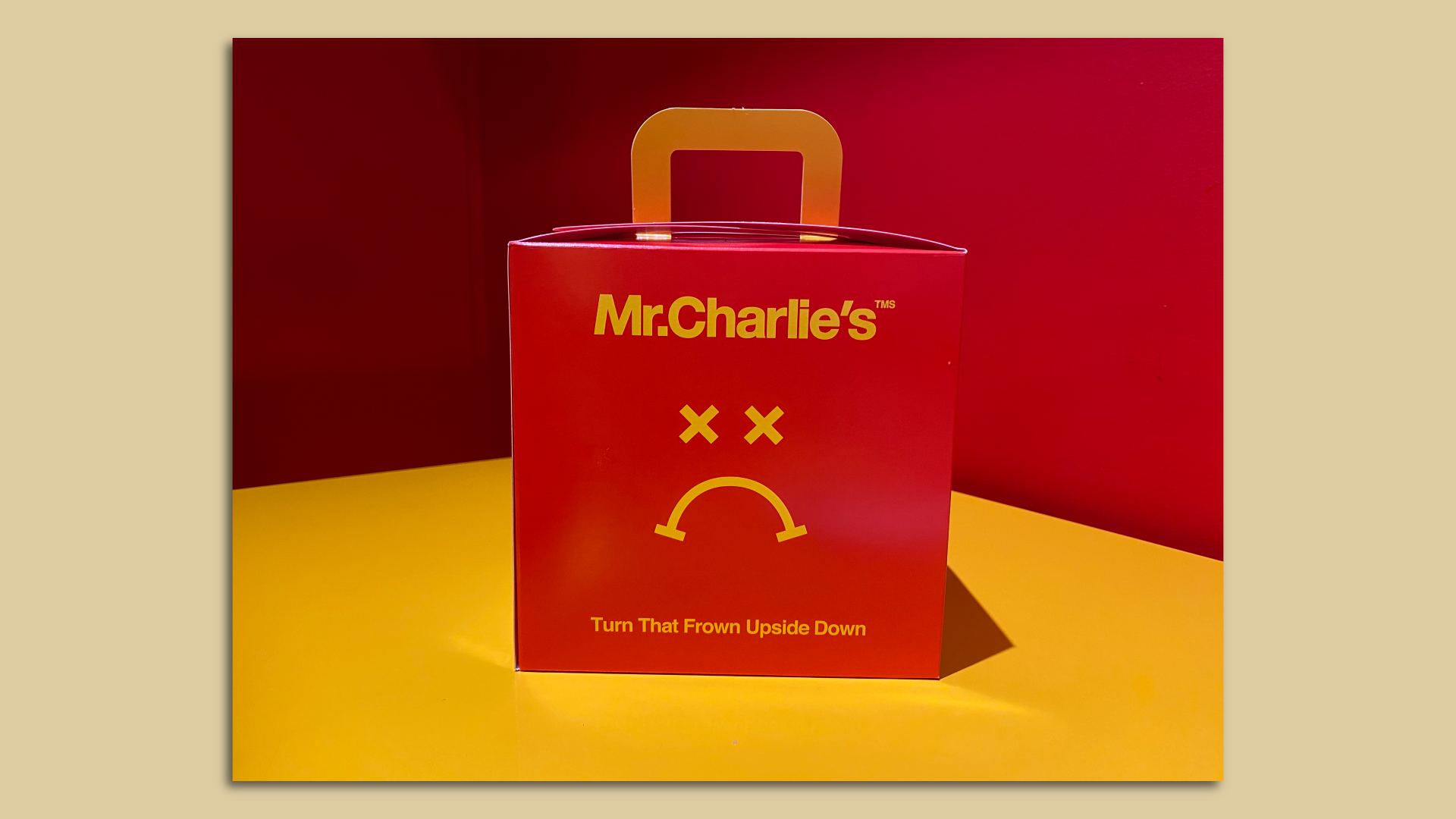 Photo of a red to-go fast food box that says "Mr. Charlie's" in yellow at top with a yellow frowny face below and the words "Turn that Frown Upside Down" sitting on a yellow table.
