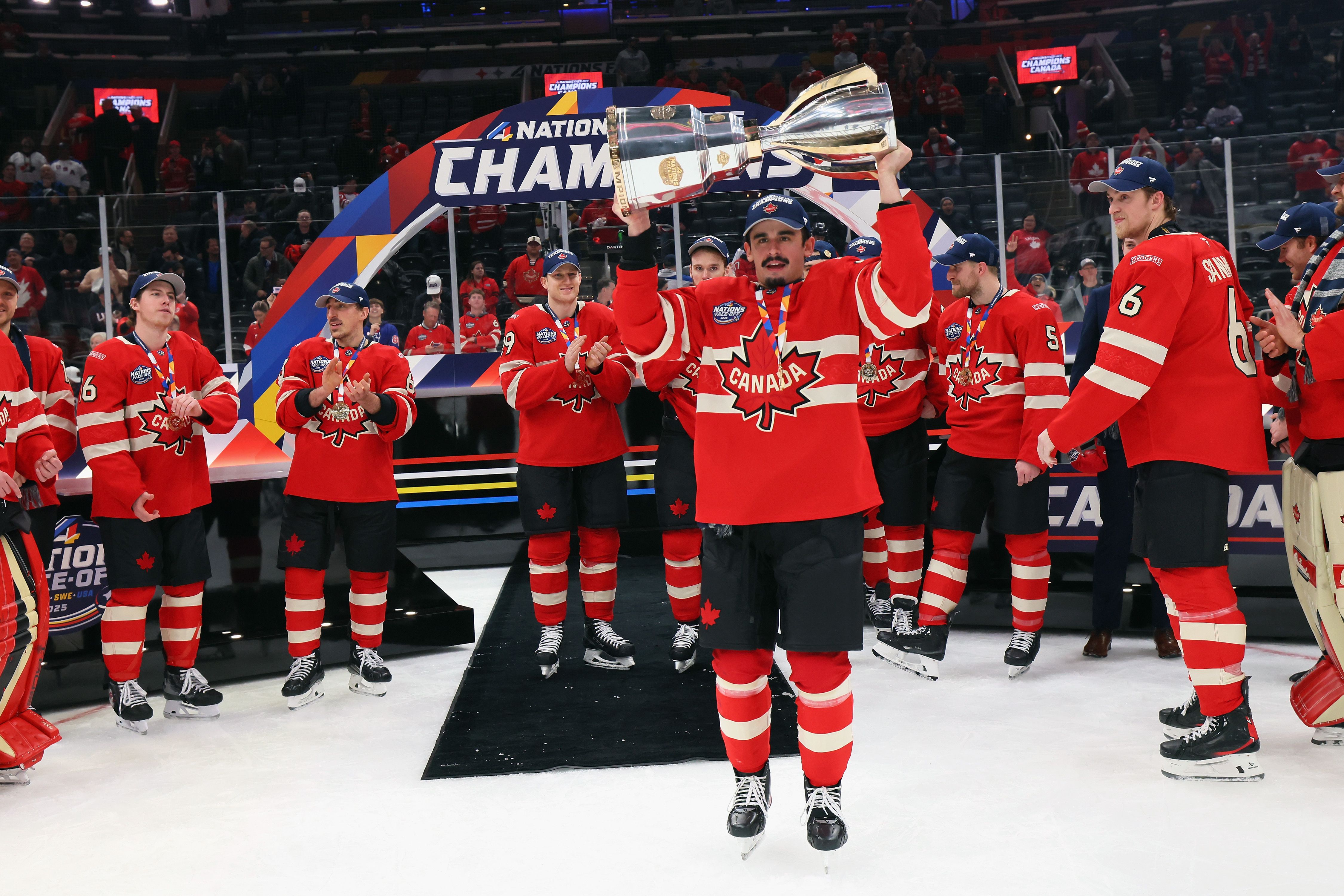 Team Canada hockey players celebrate a championship victory as one player lifts a large silver trophy while teammates in red jerseys applaud on the ice.