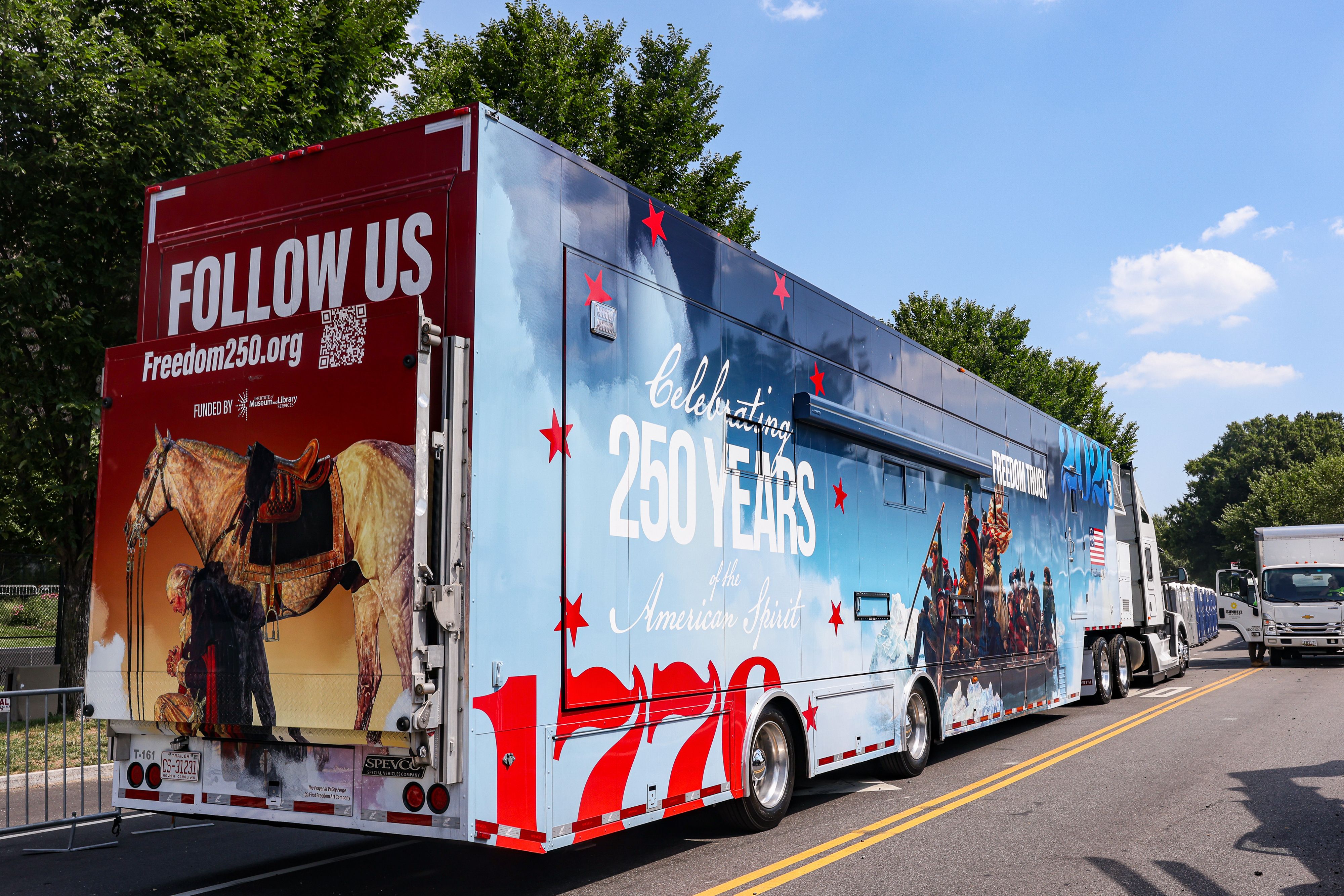 Wide promotional trailer in red and blue parked on a sunny street. The rear panel reads "FOLLOW US" with a QR code and a horse image; the blue side shows stars and large "250 YEARS" text.