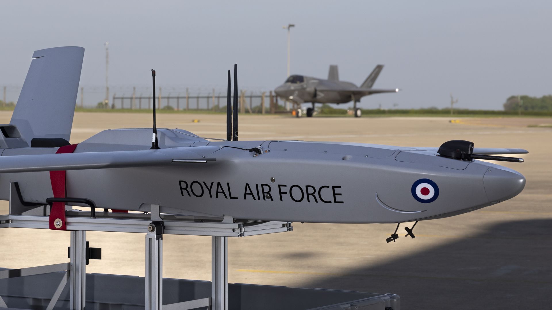 A Royal Air Force-branded drone sits on tarmac. An F-35 is seen in the background. The sky is grey.