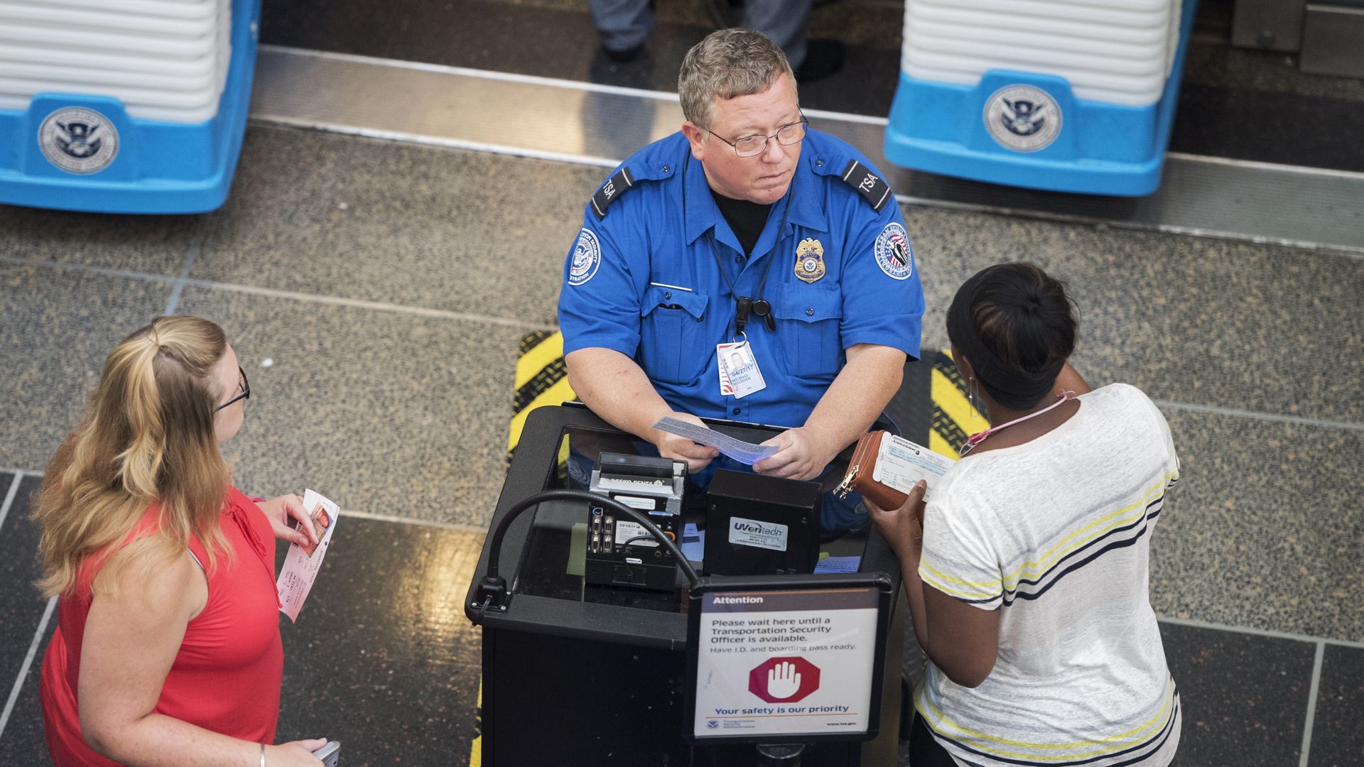 In this image, a TSA officer sits and speaks to a passenger in a white shirt.