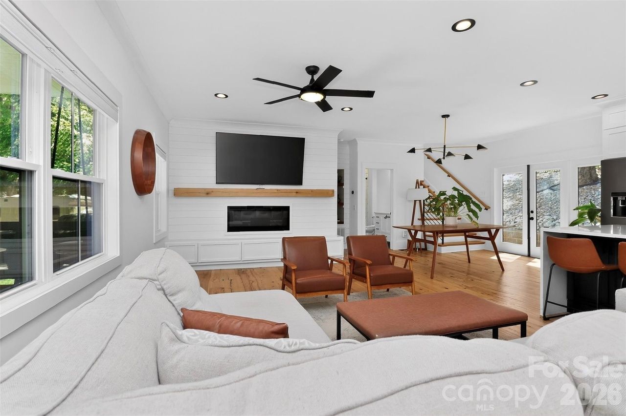 Bright open-concept living room: white walls, large windows, gray sofa with brown cushions, two brown armchairs, brown ottoman, fireplace with mounted TV, wooden staircase, and a dining area near the kitchen.
