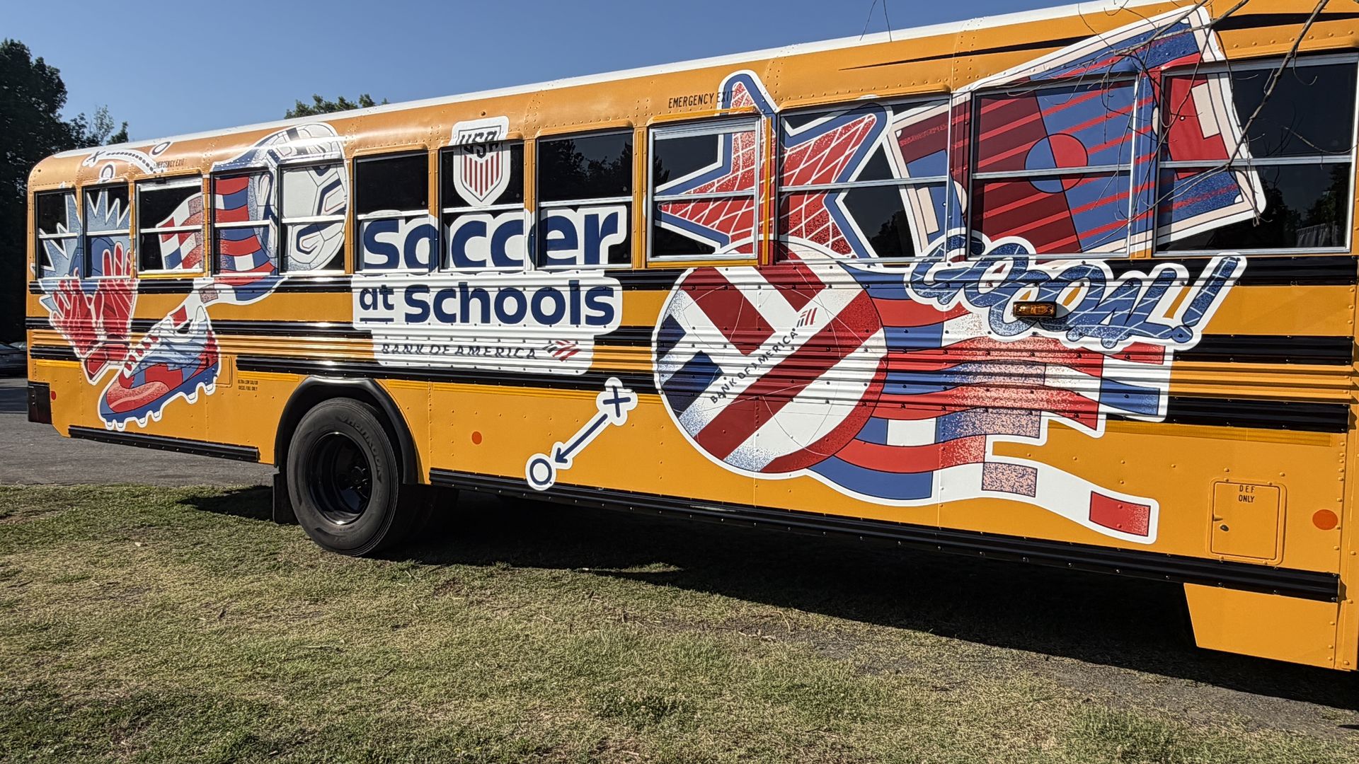 Bright yellow school bus covered in bold graffiti art, featuring the blue text "soccer at schools" amid red, white, and blue shapes, parked on grass under a clear blue sky.