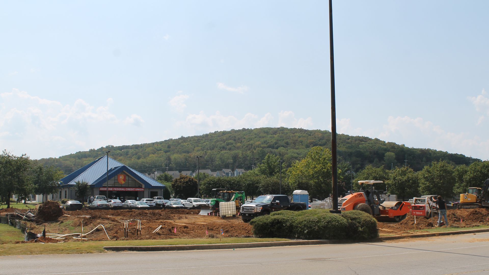 Construction site with heavy machinery and workers digging beside a road; a Redstone Federal Credit Union building with blue roof and parked cars is in the background against a green hill under a blue sky.