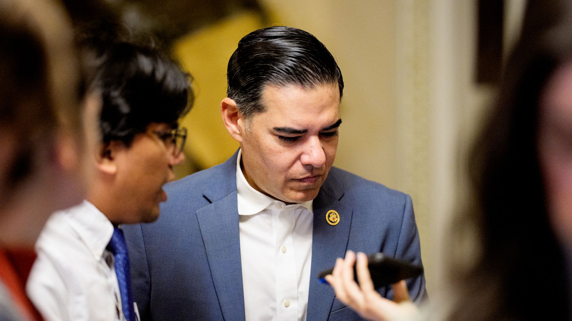 Robert Garcia, wearing a blue suit, speaking to reporters in front of a white and yellow wall.