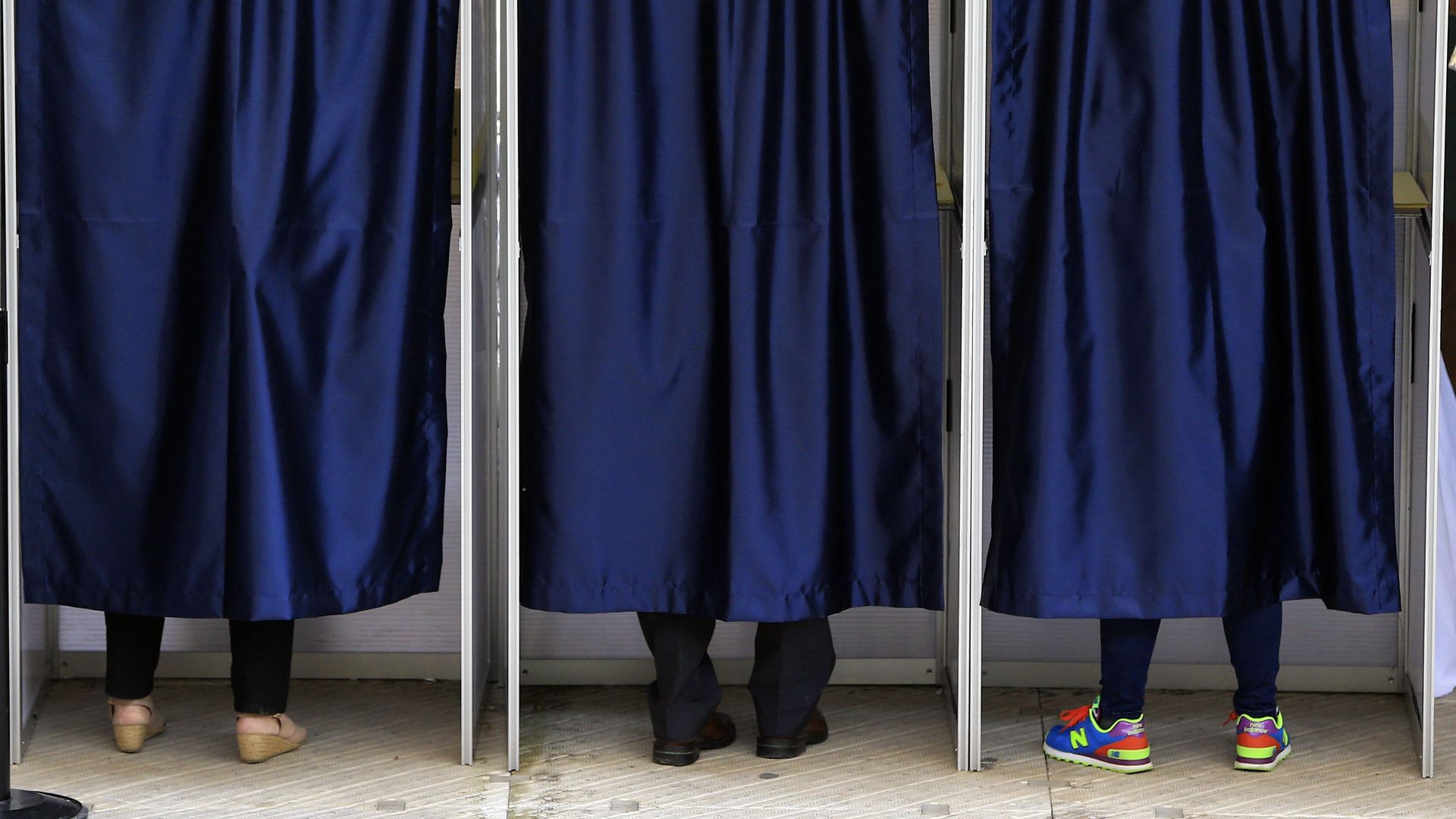 Blue curtains covering three voting booths. You can see the shoes of three different people at the bottom of each curtain