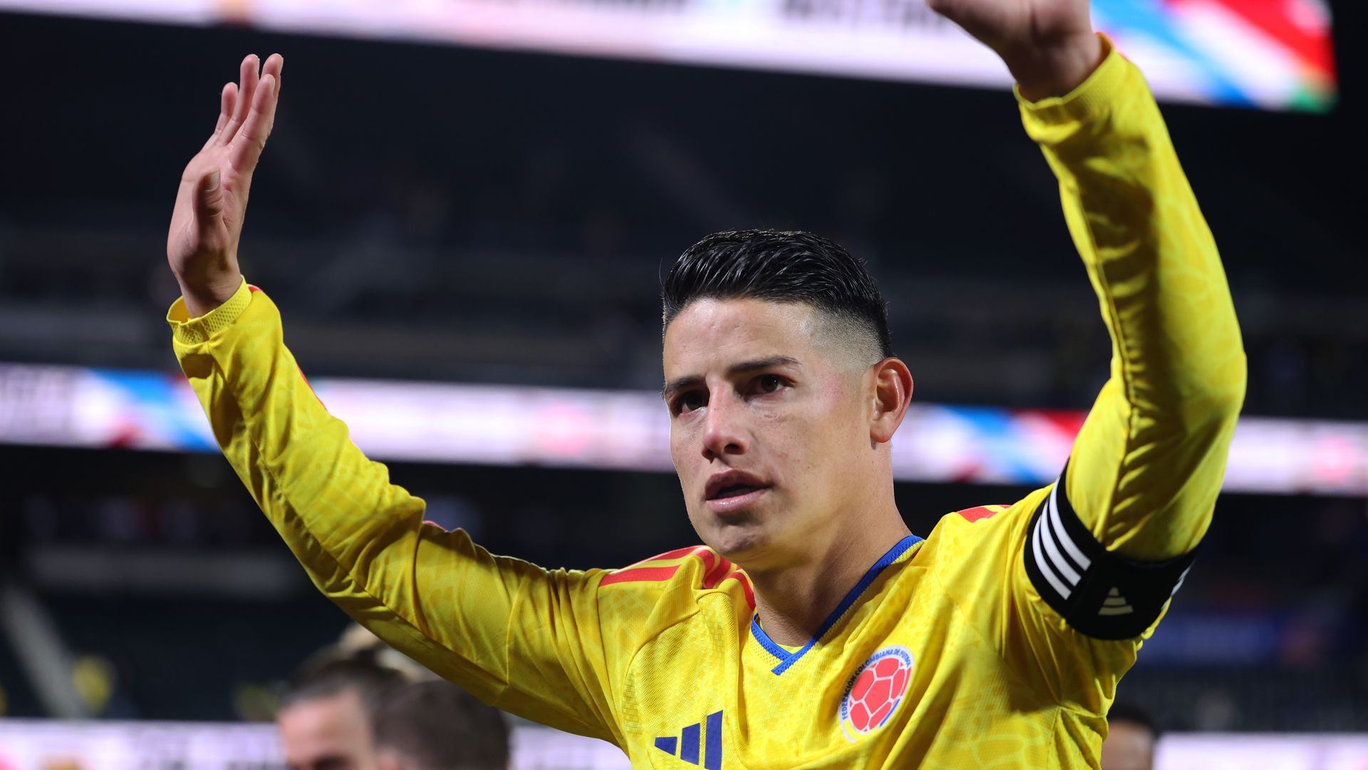 James Rodriguez wears a yellow jersey and waves his hands in the air. 