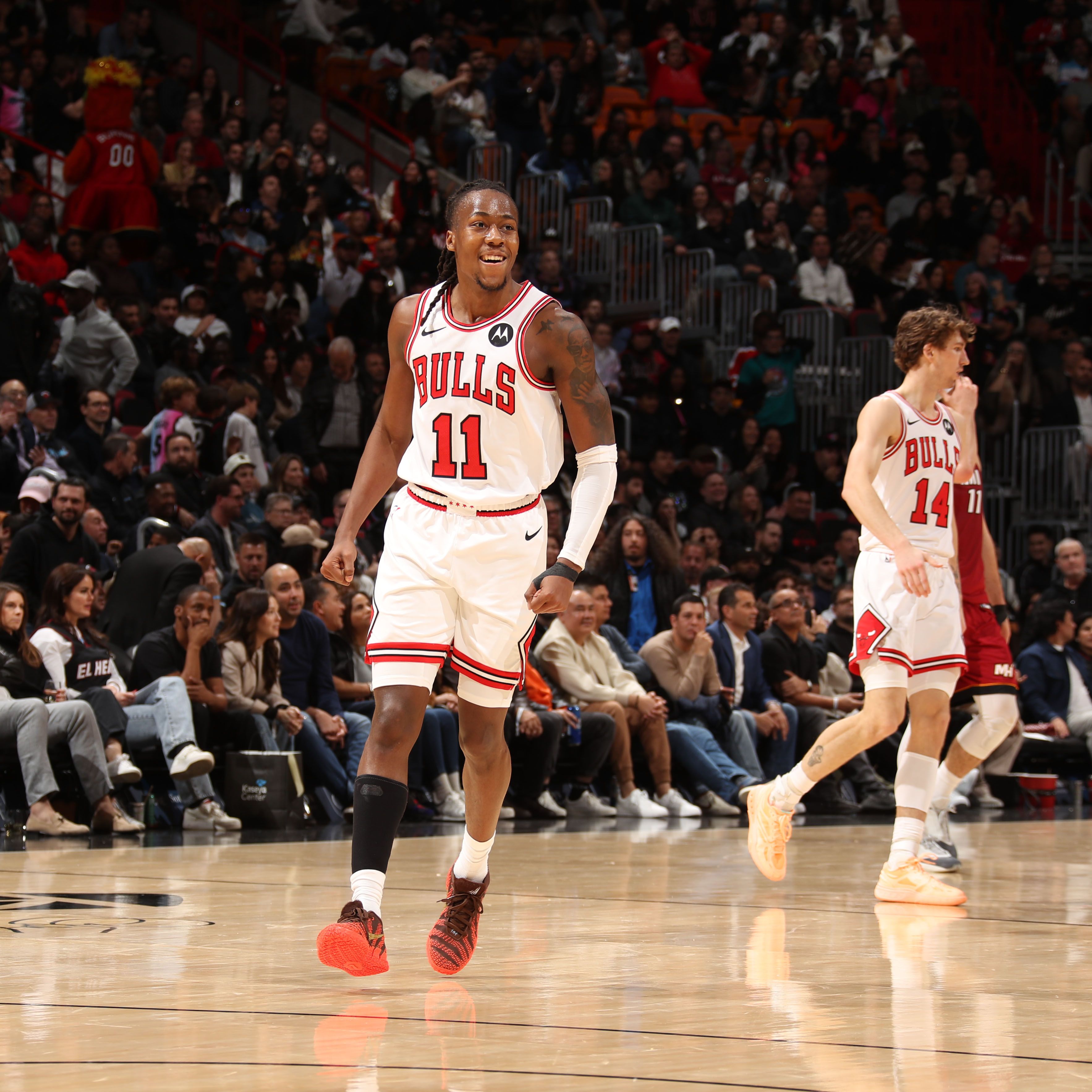 Two Chicago Bulls basketball players on court during a game, wearing white uniforms with red and black trim, with a crowd in the background including a mascot dressed in red.