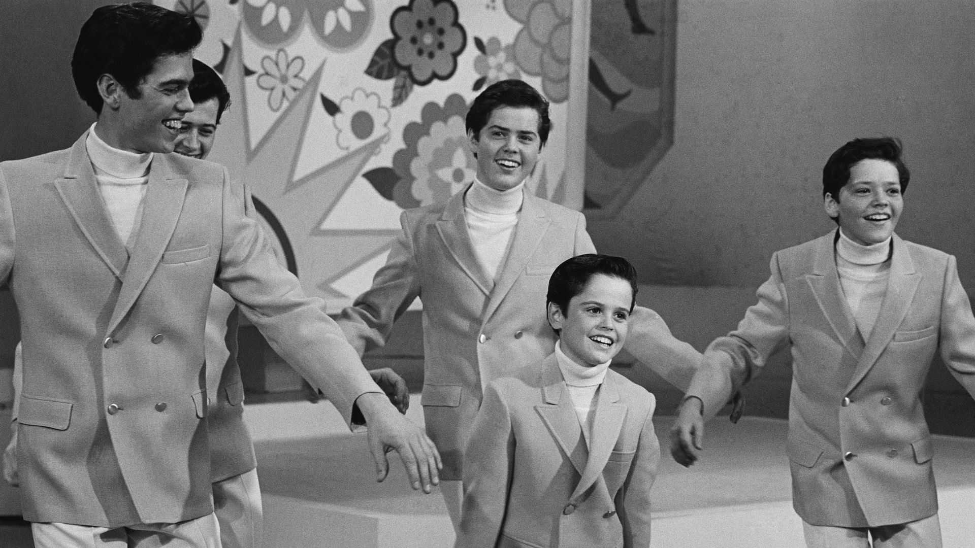 A black and white photo of five boys dancing in dress coats on a 60s TV show.