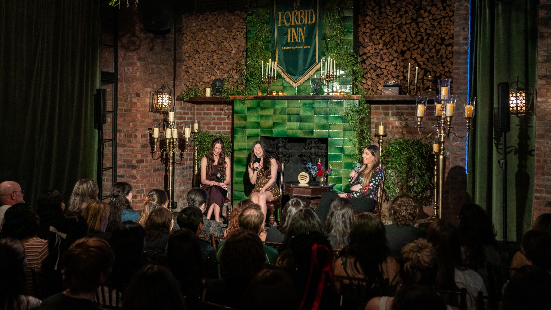 Three women seated on stage in front of a green tiled fireplace with "The Forbid-Inn" banner, speaking to a seated audience in a cozy, candlelit room with brick walls and green curtains.