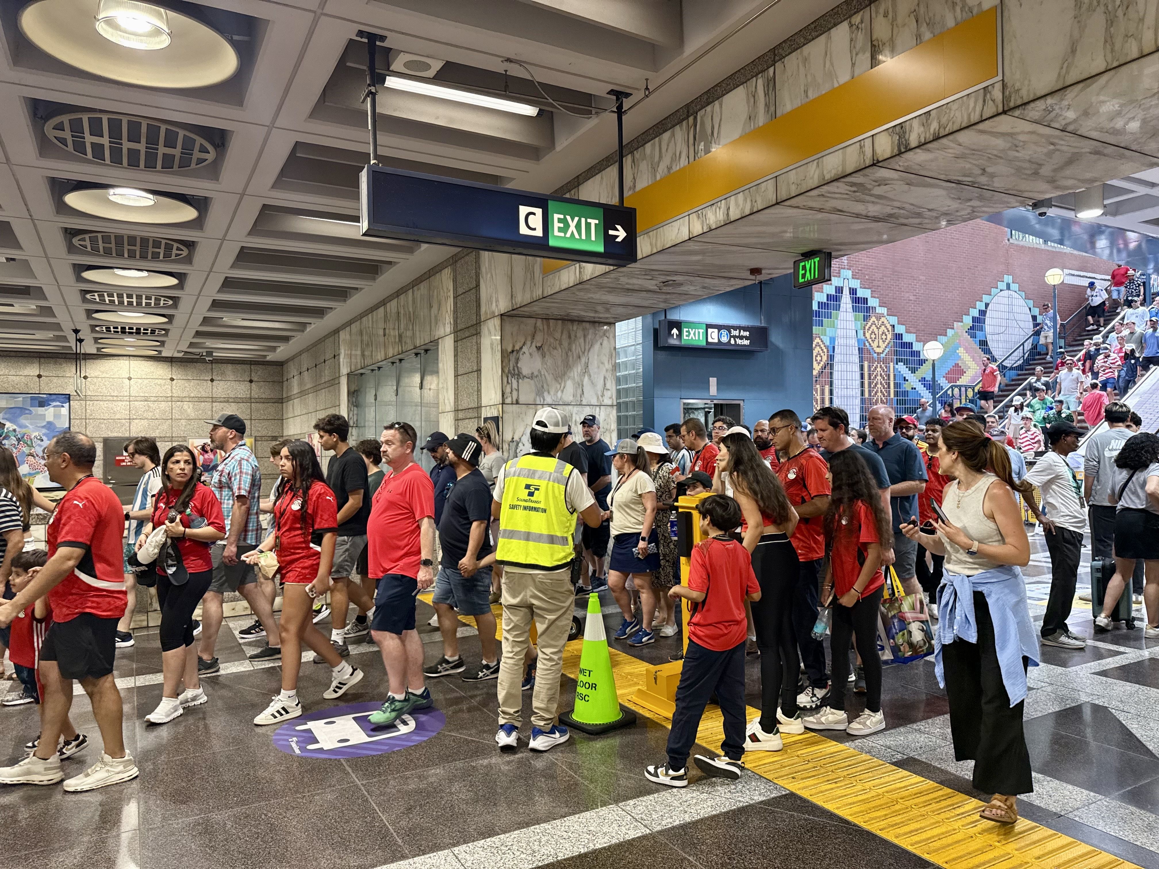 Busy subway concourse filled with a crowd of people wearing red shirts. Marble walls, ceiling lights, and green EXIT signs. A yellow tactile path runs along the floor; stairs visible to the right.