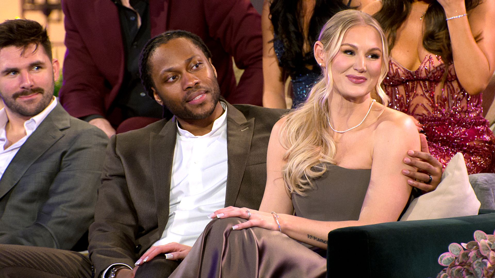 Three people seated on a sofa during a TV show: a man in a brown suit beside a blonde woman in a taupe strapless dress; others in formal wear in the background.