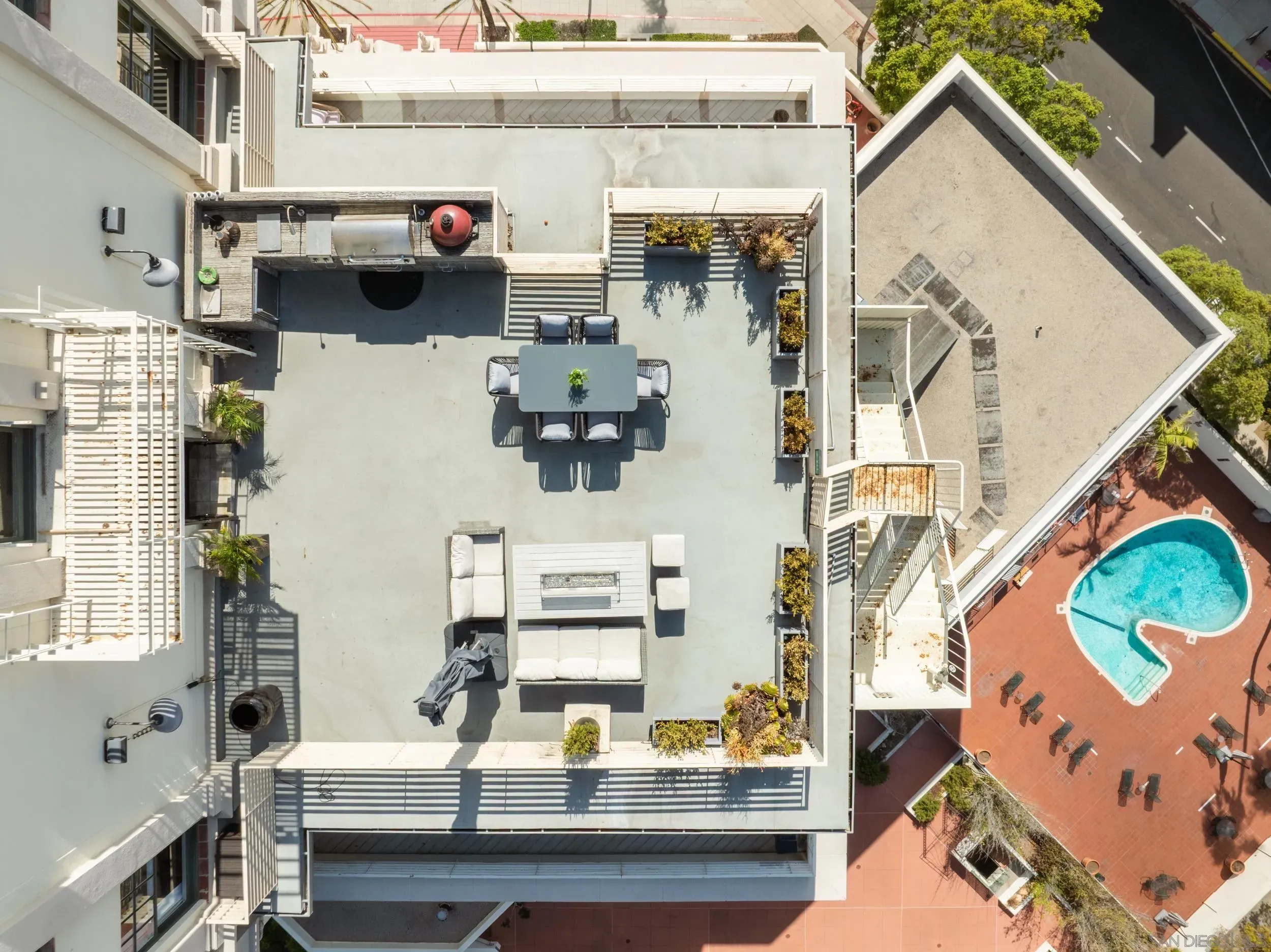 An aerial view of a modern rooftop terrace: gray concrete deck, outdoor dining table with eight chairs, planters and a grill, lounge seating, and a blue pool with red surrounding pavement.