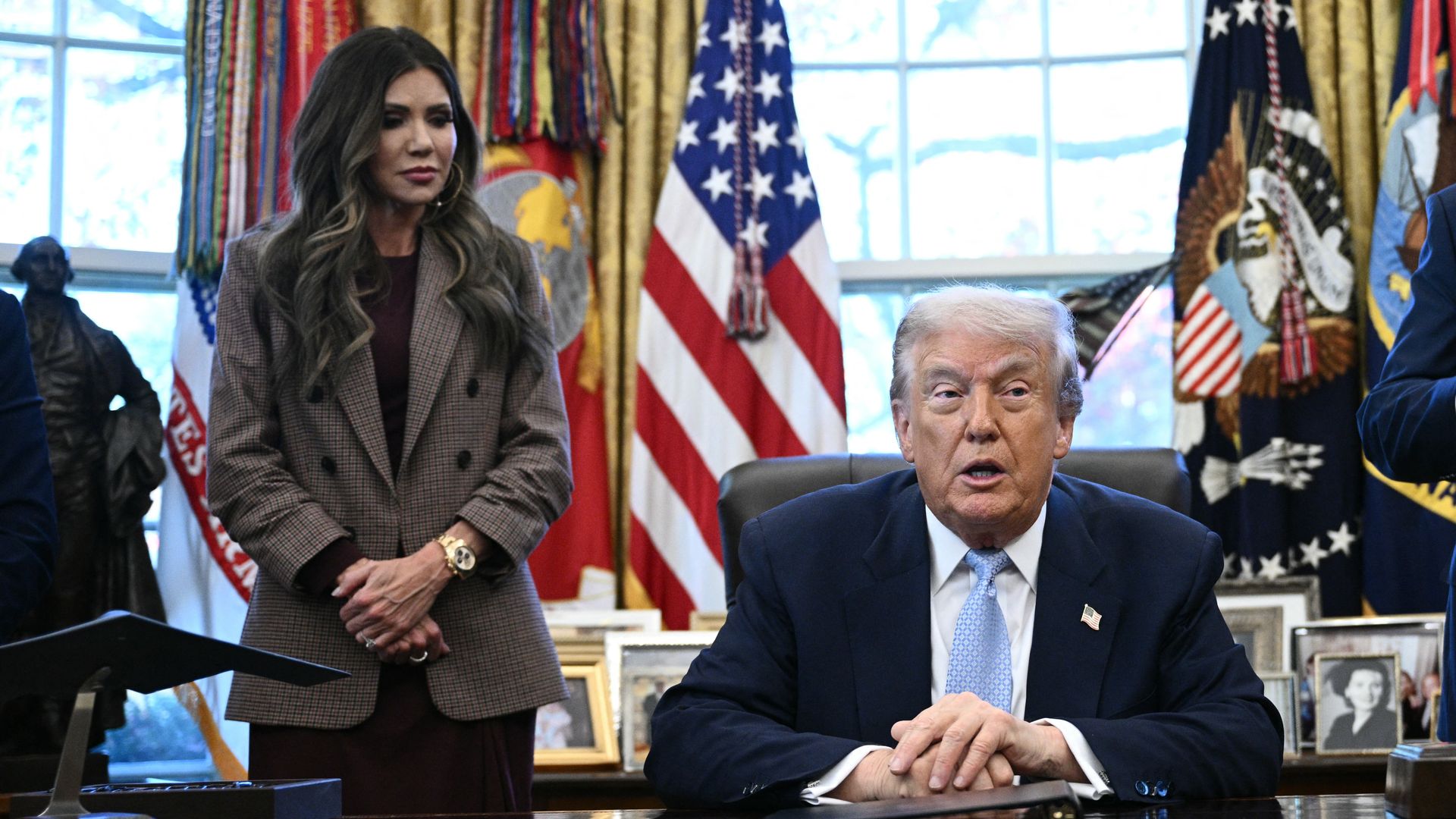 A standing, dark-haired Security Kristi Noem, wearing a brown suit jacket with a black top over a brown dress, clasps her hands and looks at President Trump, sitting at a brown desk in the White House, wearing a navy jacket, white shirt and blue tie, clasping his hands while speaking.