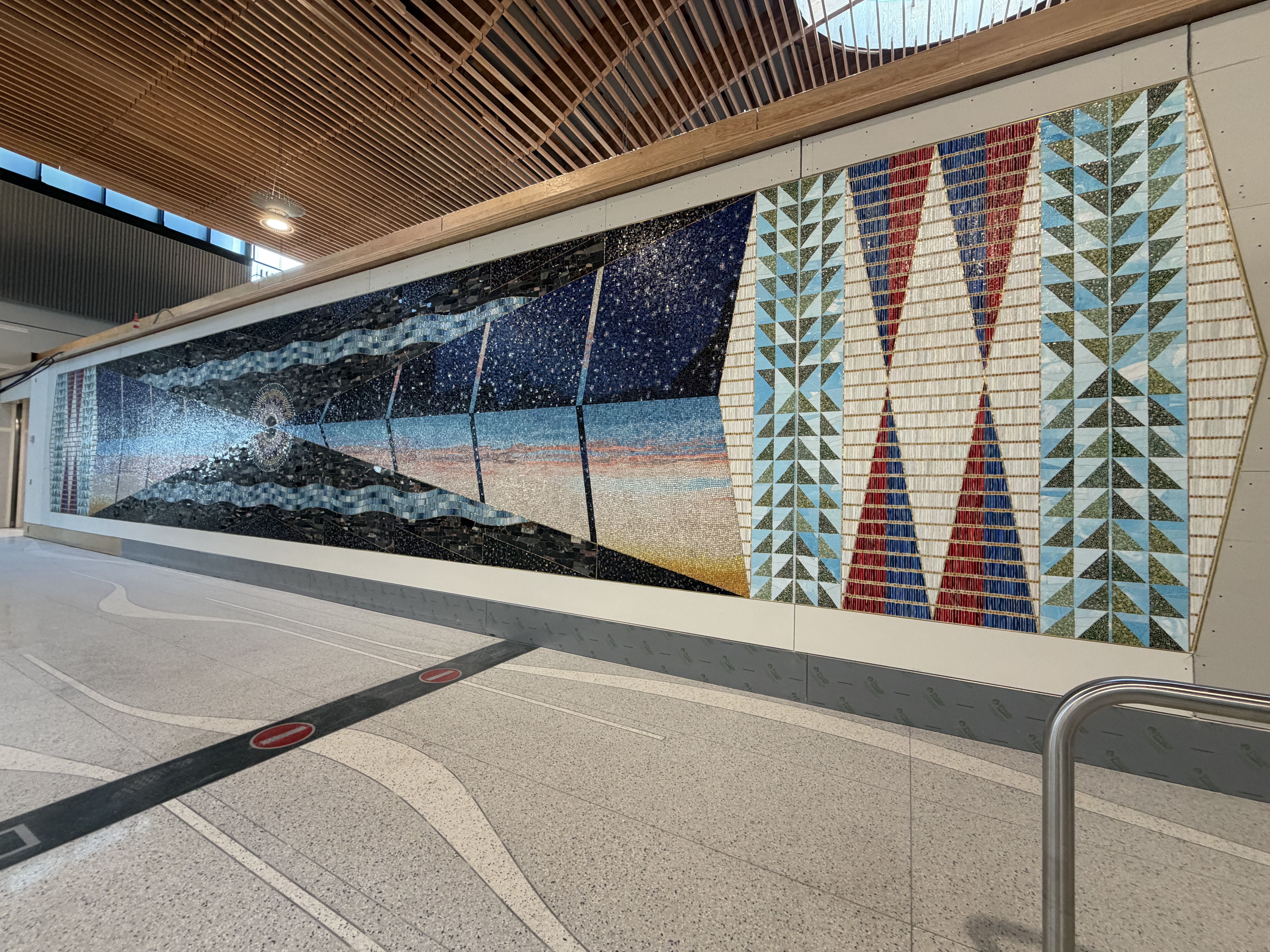 Long mosaic mural with abstract geometric shapes in blue, red, white, and green, framed by light wall panels, indoors with wooden ceiling and terrazzo floor.