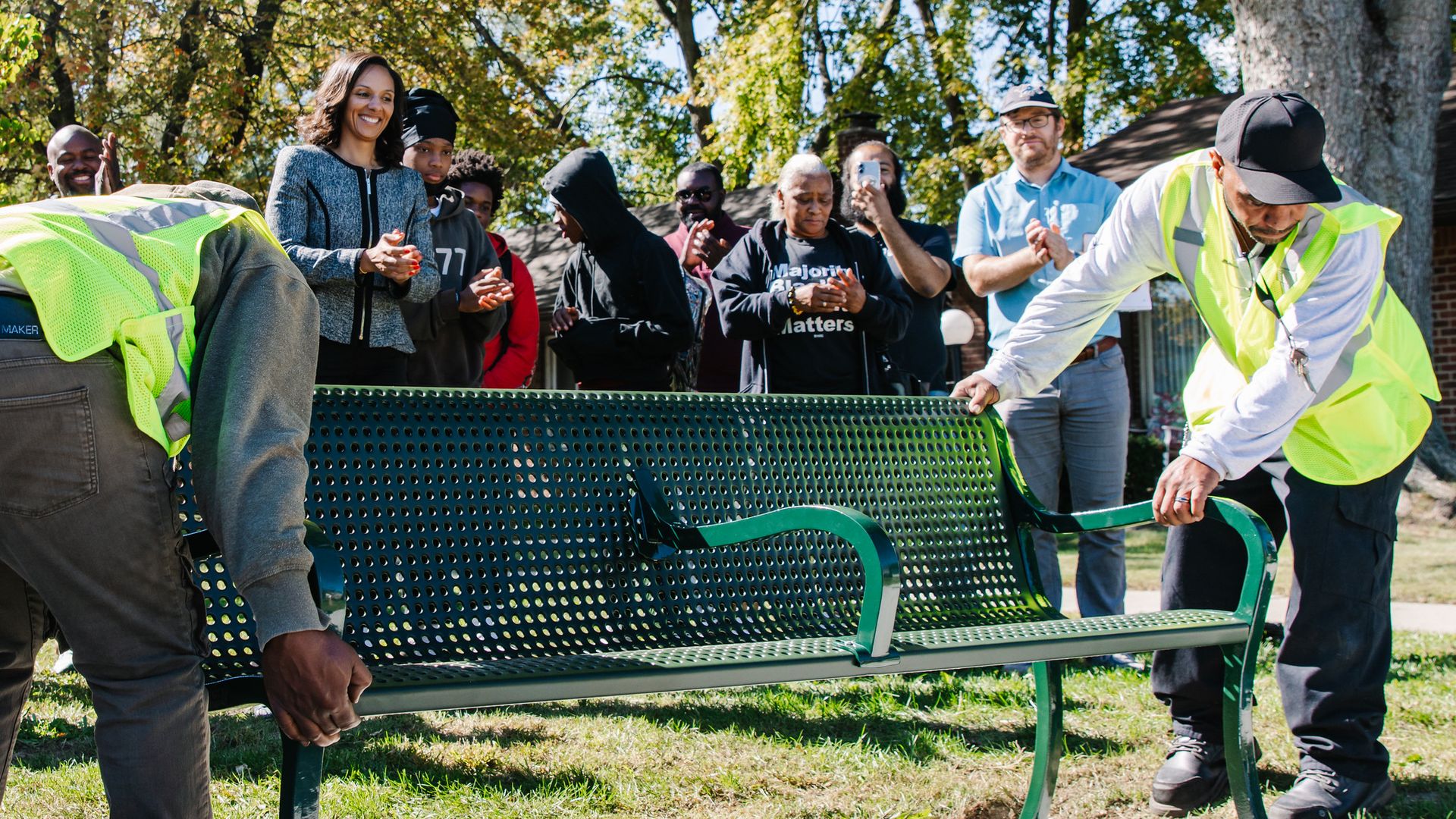 A group of people outdoors near a green perforated bench, with workers installing it; Mary Sheffield stands center as others clap, with trees in the background.