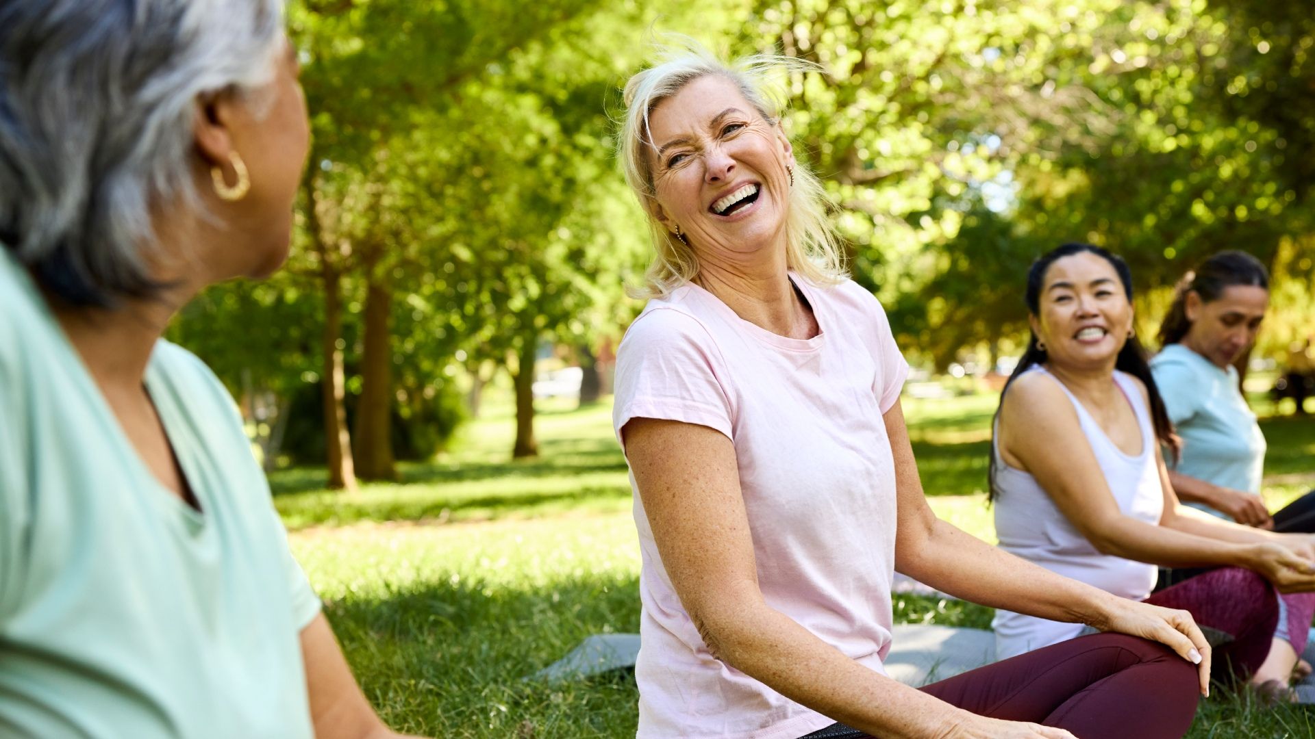 People doing outdoor yoga in a sunlit park. In the center, a smiling blonde woman in a pink shirt laughs as others in pastel tops exercise on mats nearby, with green trees in background.