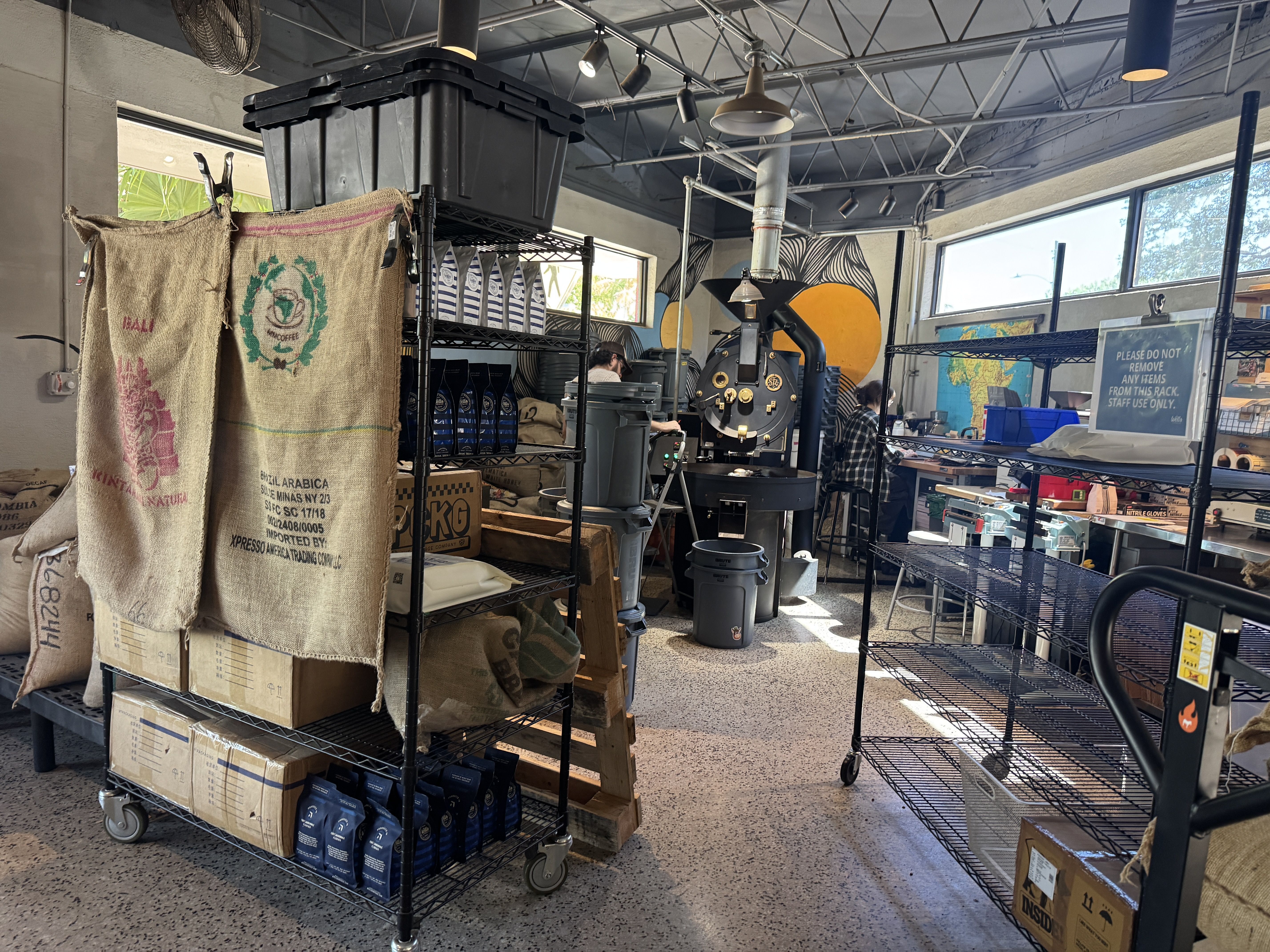 Industrial roastery interior with burlap coffee bags on a cart, metal shelving, a large coffee roaster, exposed pipes and hanging lights; two people work in the back; sign visible on the right.