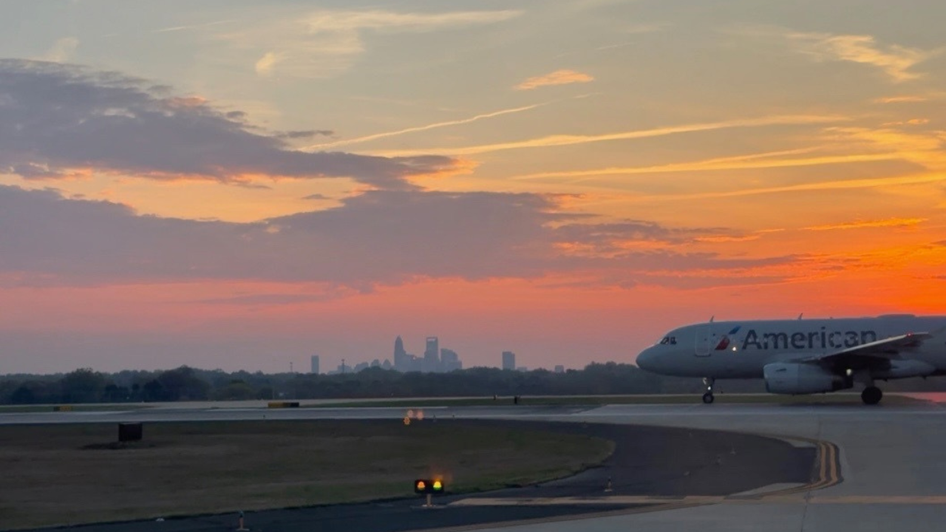 An American Airlines jet taxis along a runway at sunset, with an orange-pink sky and a distant city skyline on the horizon.