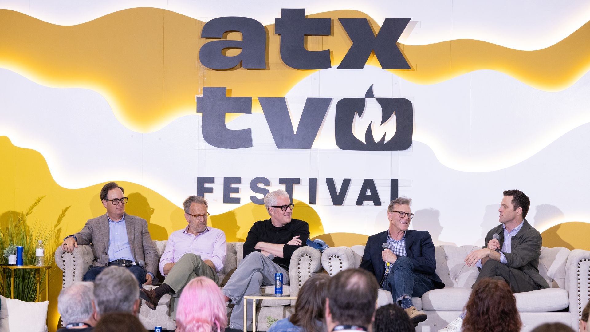 Panel discussion on a stage at the ATX TV Festival; a yellow wavy backdrop with large signage reading ATX TV FESTIVAL. Five men sit on a couch, microphones in hand, audience visible in the foreground.