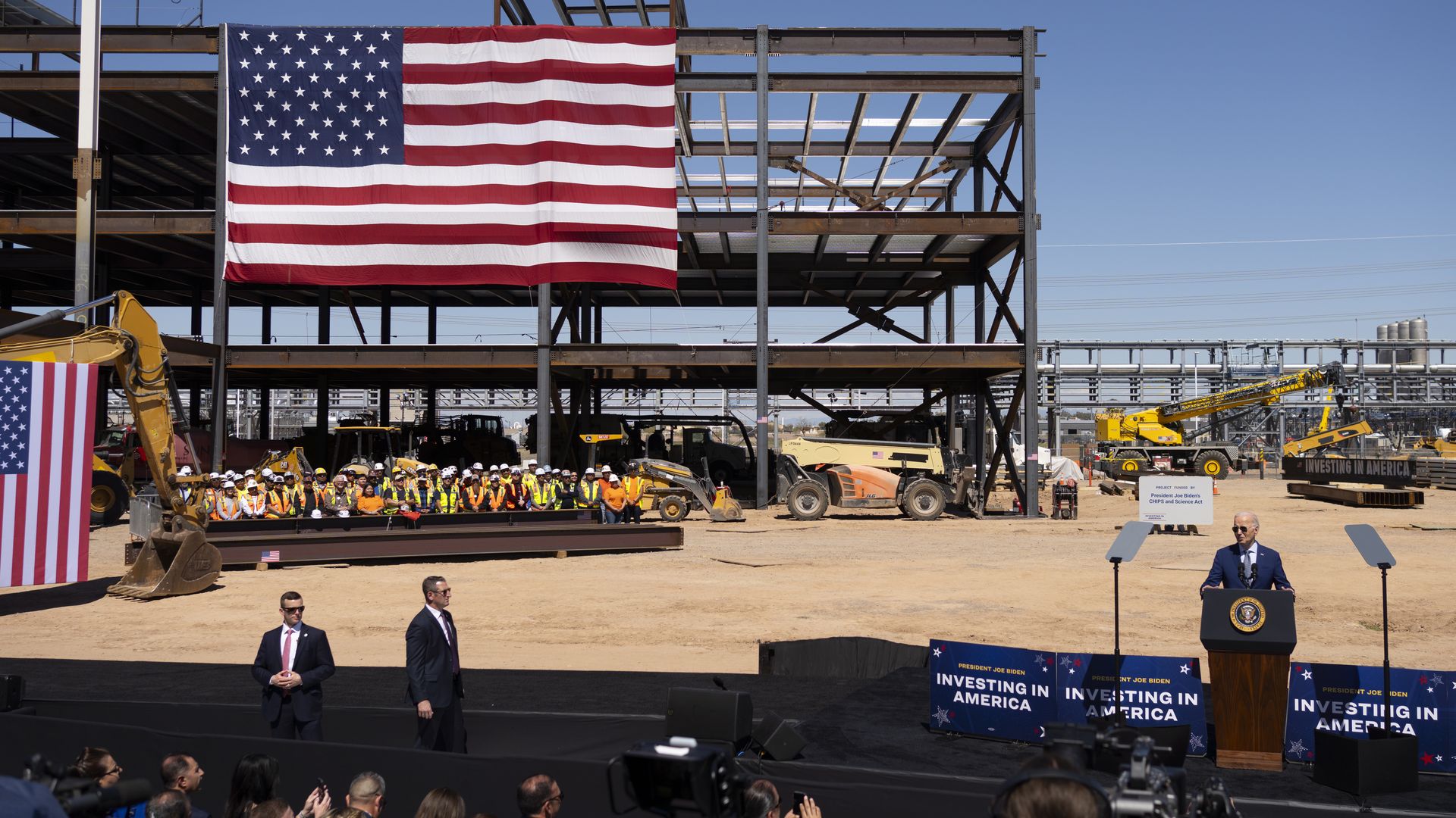 President Biden speaking on a stage at a construction site with an American flag in the background. 