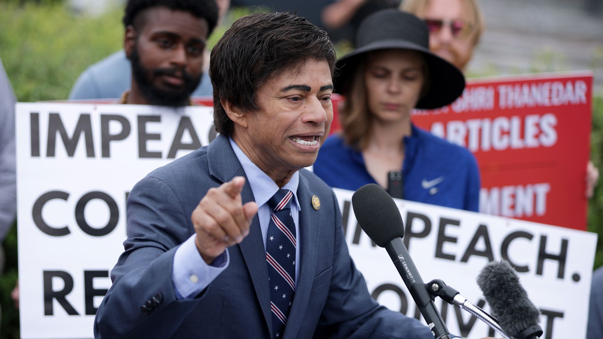 Rep. Shri Thanedar wearing a blue suit and speaking in front of people wearing signs.