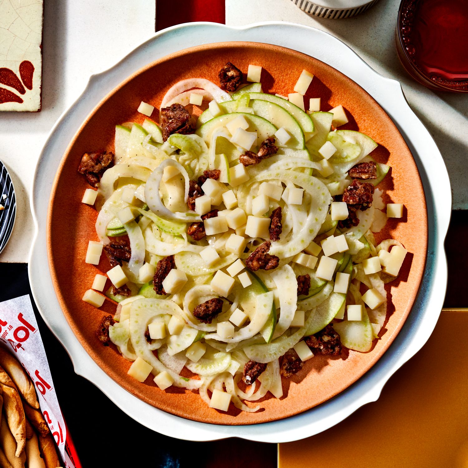 Salad with thinly sliced green apple, fennel, cubed cheese, and candied nuts on an orange plate with a white rim on a tiled surface.