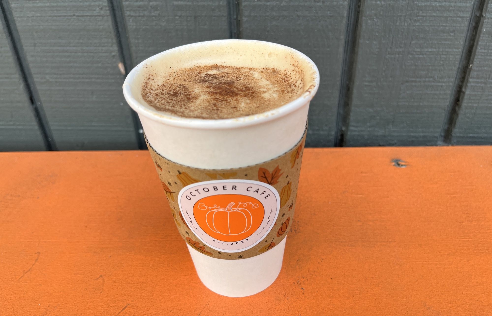 A to-go coffee cup with cinnamon dusting on the foam sits on an orange table. Its sleeve has an orange pumpkin logo and autumn graphics, with a dark gray wooden fence behind.