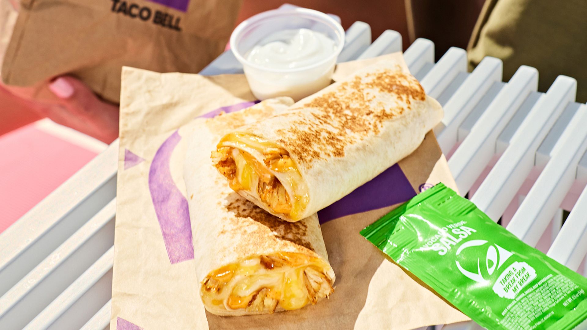 Two burritos filled with melted cheese and chicken sit on parchment on a light blue bench, with a cup of sauce, a green salsa packet, and a Taco Bell bag in the background.