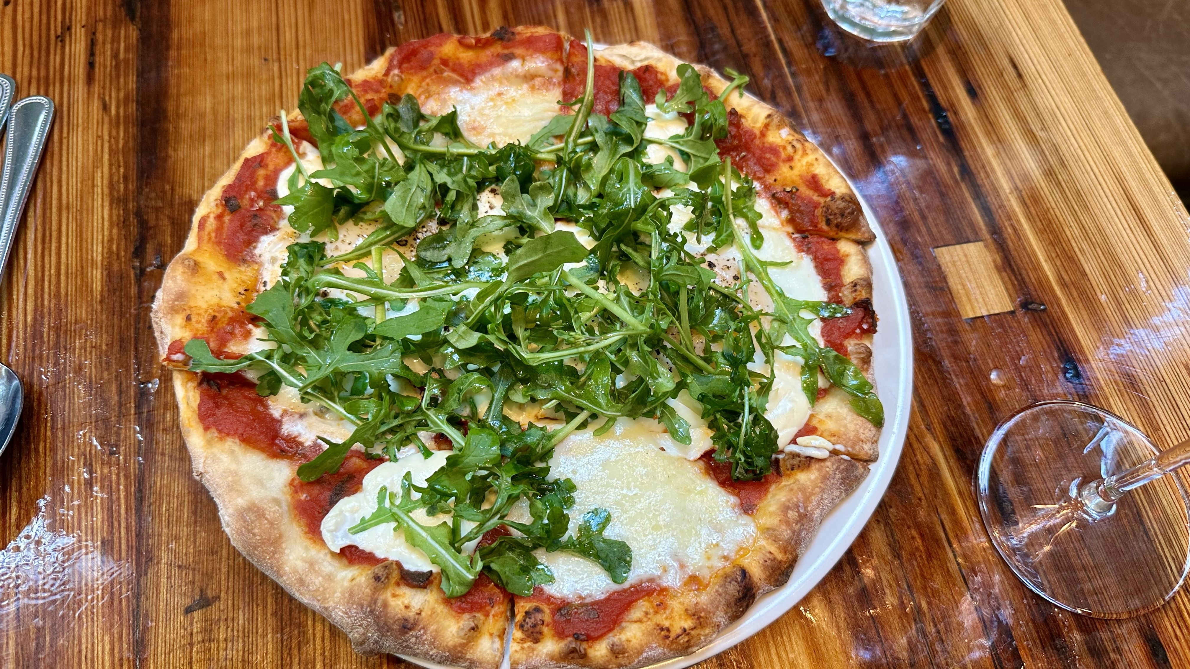 Top-down view of a pizza with tomato sauce, melted cheese, and a large arugula topping on a white plate, placed on a wooden table with cutlery and a glass nearby.