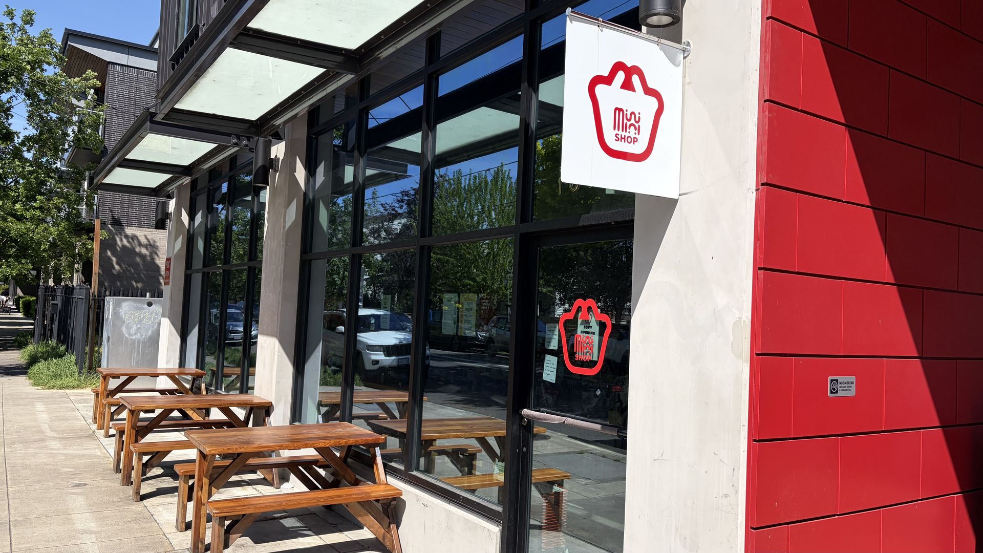 Storefront with large glass windows, a bold red wall on the right, and outdoor wooden picnic tables along the sidewalk. A white hanging sign shows a red bag icon and MINI SHOP.