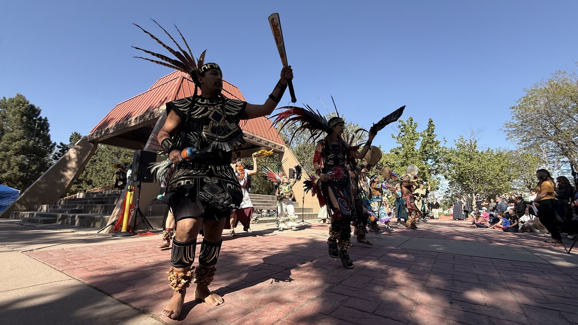 Indigenous dancers in black regalia with tall feather headdresses perform on a plaza, wielding a carved staff and fans; audience sits to the right as a red-roofed building and trees frame the view.