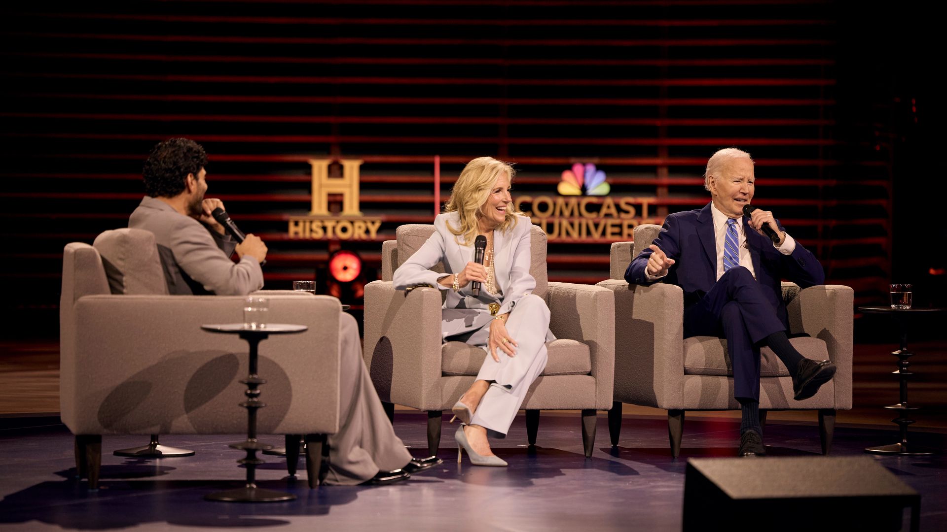 Three panelists sit on a stage in gray and blue suits, each holding a microphone, engaged in discussion. A History sign and NBCUniversal logo glow against the red-lit backdrop.