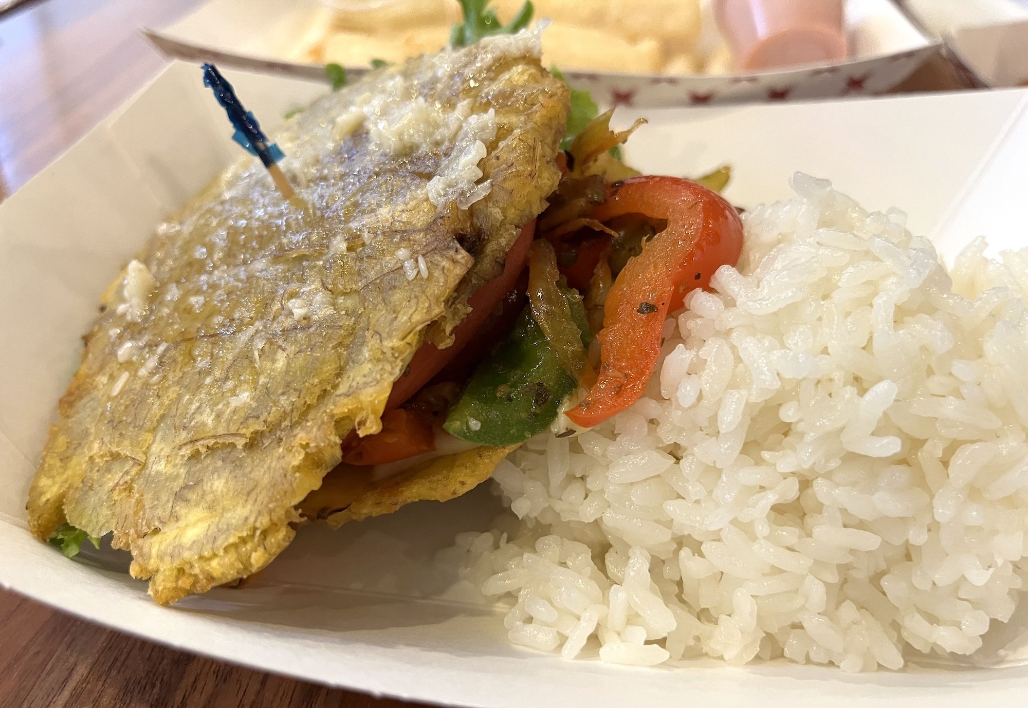 Golden fried fish fillet laid over white rice with red and green peppers in a white takeout box.
