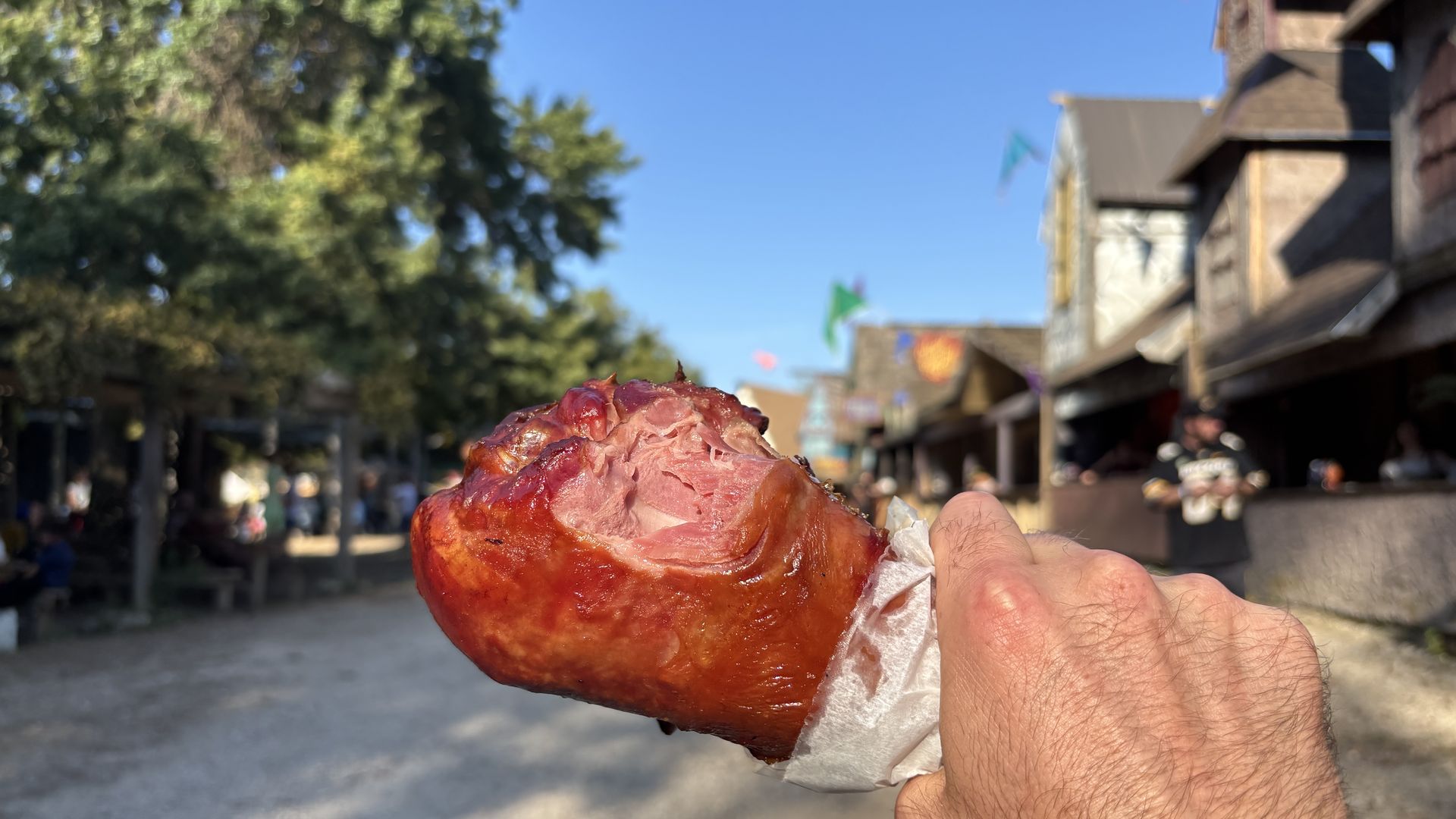 Close-up of a glazed reddish-brown roasted meat held in a napkin by a hand, outdoors on a sunny street with green trees and wooden buildings in the background.