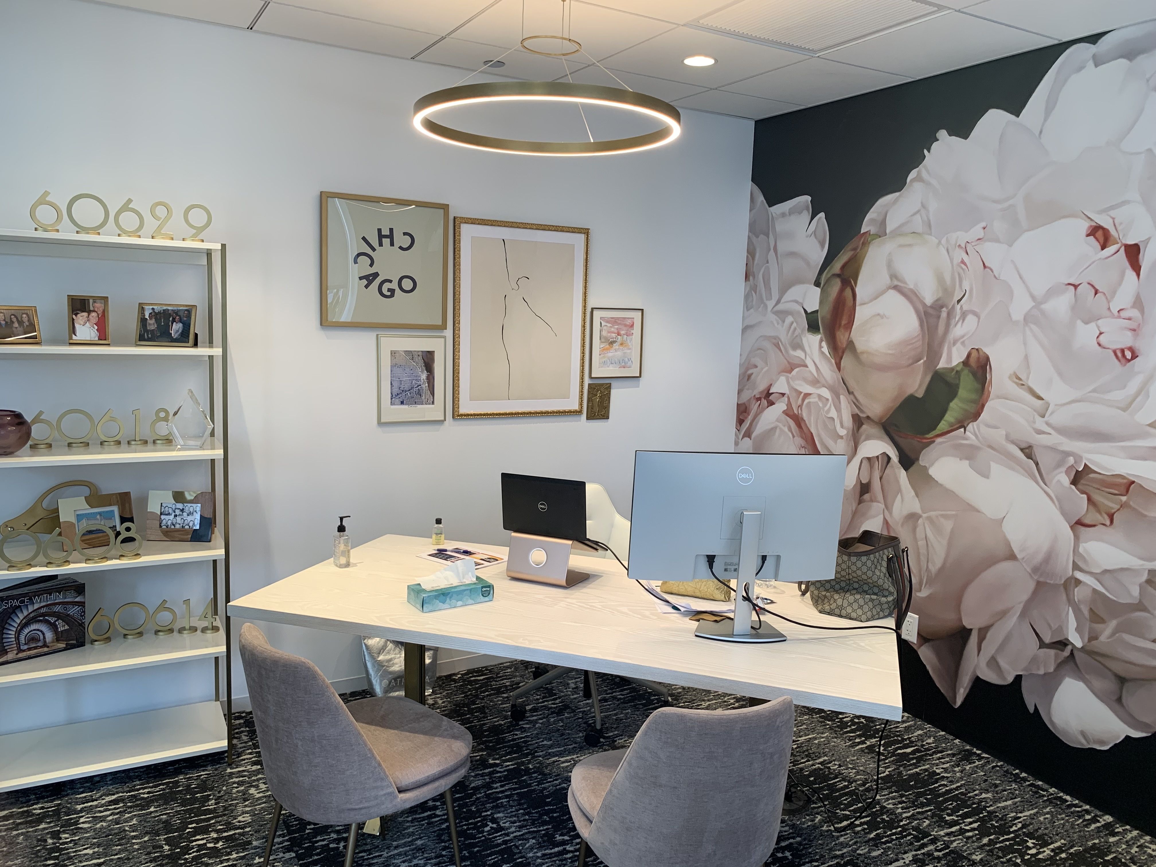 Modern office with white desk, two gray chairs, a Dell computer and laptop. Large floral mural wallpaper on right wall. Shelves with photo frames and gold numbers, framed abstract art on white wall.