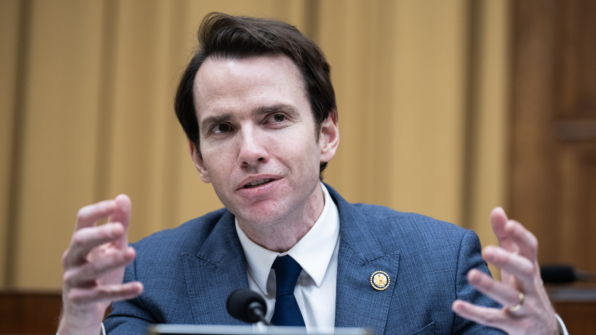 A man in a blue suit and dark tie gestures with hands while speaking at a formal setting, seated behind a nameplate labeled "Mr. Kiley," with a microphone in front of him.