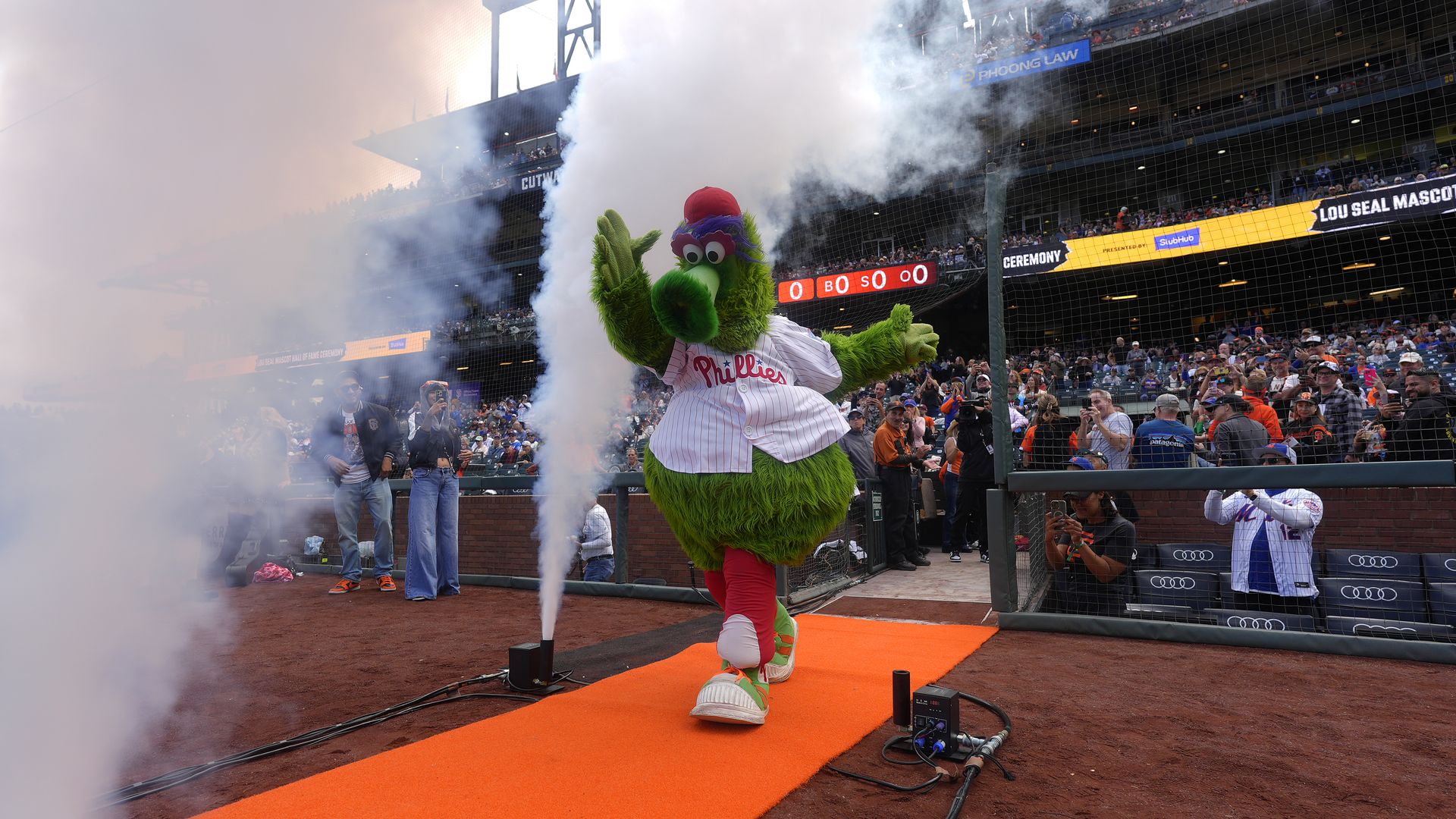 The Phillie Phanatic mascot in a white Phillies jersey walks on an orange carpet with smoke effects at a baseball stadium, with fans in the stands and around the field.
