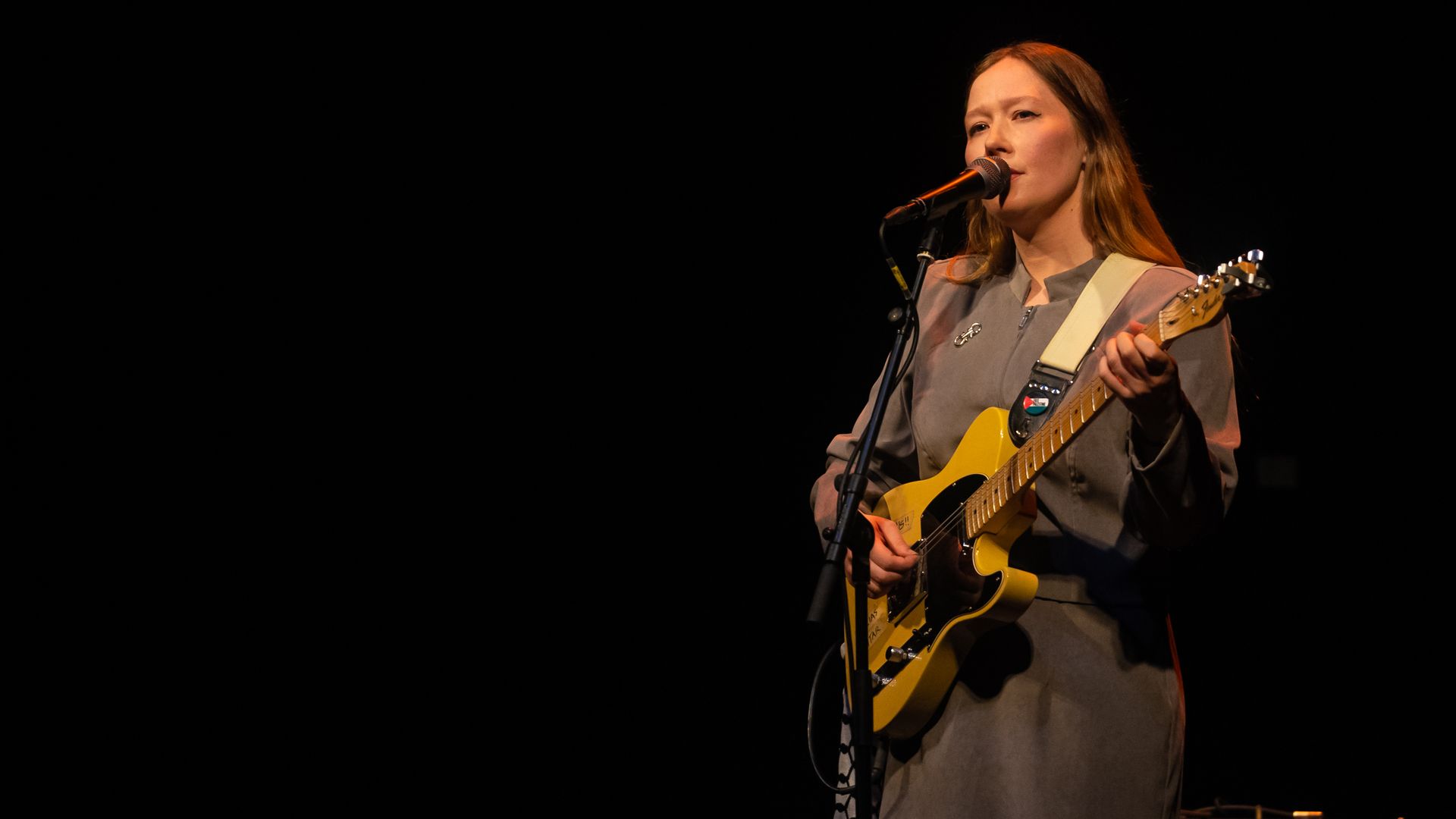 Female musician on stage sings into a microphone while playing a yellow electric guitar; warm lighting, dark background, and an amplifier nearby.