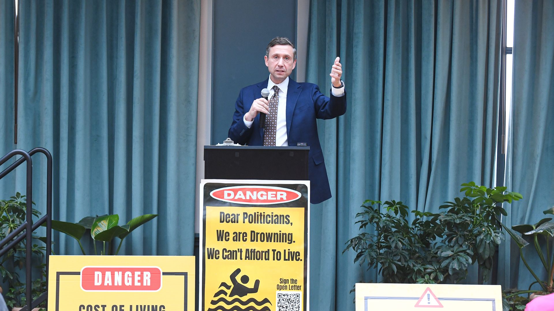 DNC chair Ken Martin, wearing a blue suit and holding a microphone while speaking at a podium with a mock yellow "Danger, drowning" sign on it, in front of a teal curtain.
