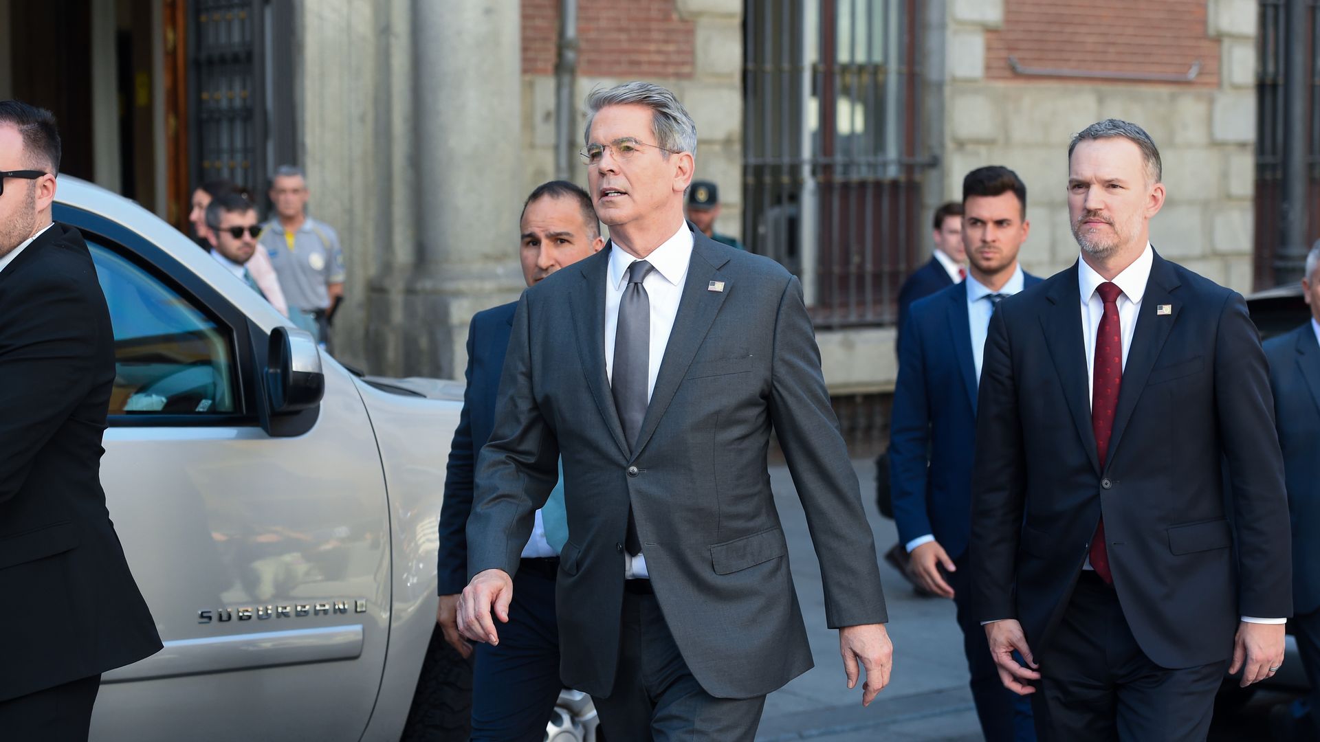 A group of men in suits walking from right to left away from a building and in front of an SUV. 