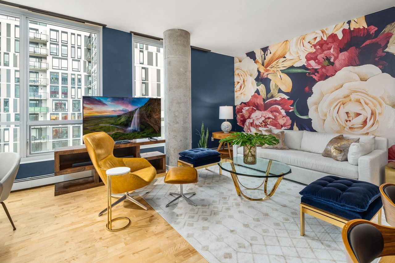 A living room with a colorful floral mural and large windows facing an apartment building.