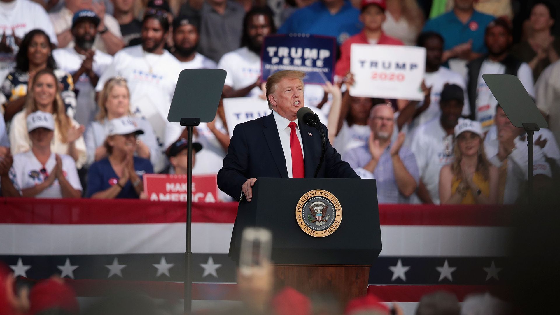 In this image, Trump stands at a podium and speaks into a microphone with a crowd of supporters in rows behind him.